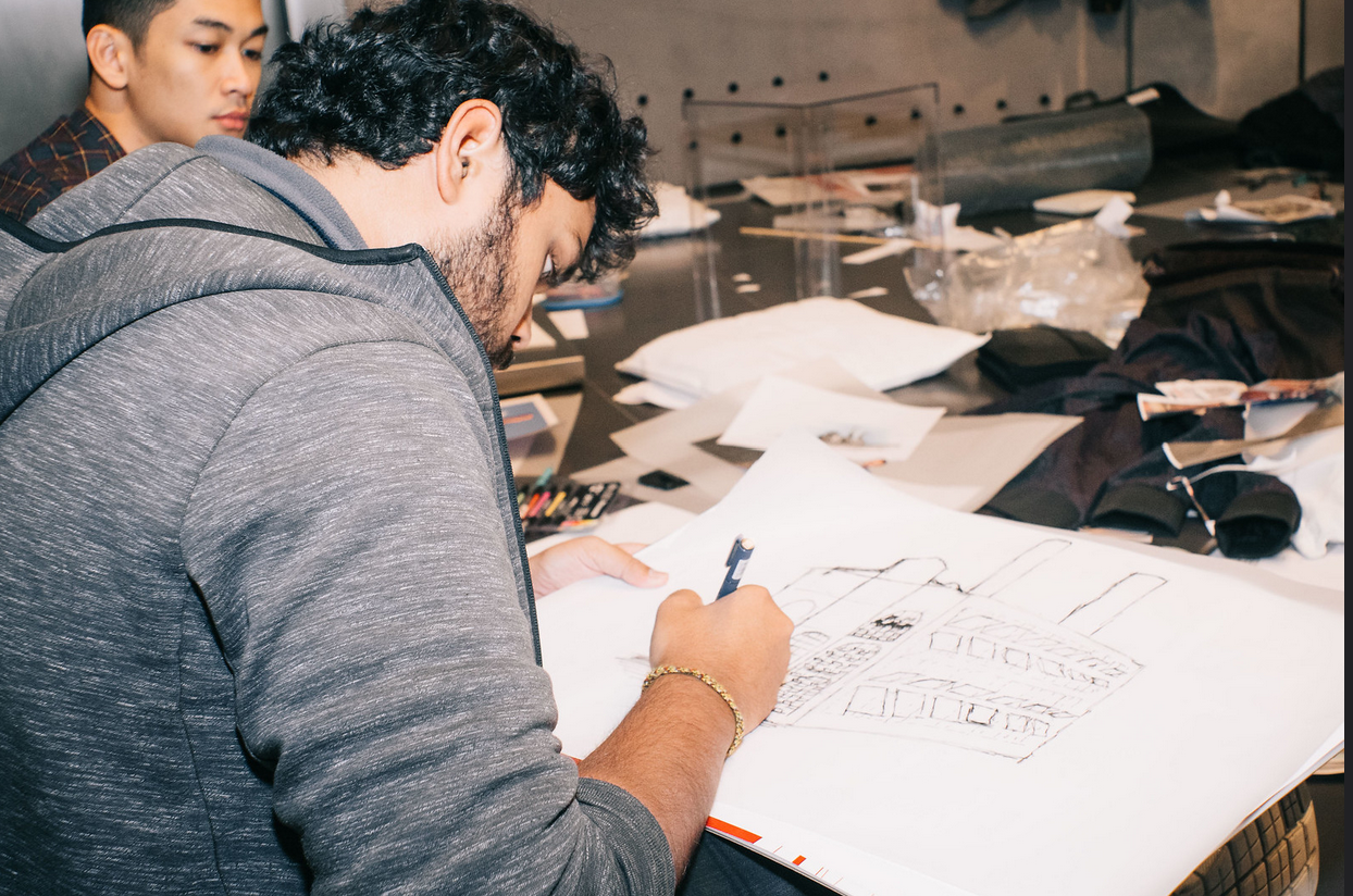 Person sketching a building blueprint on a large sheet of paper at a cluttered worktable.