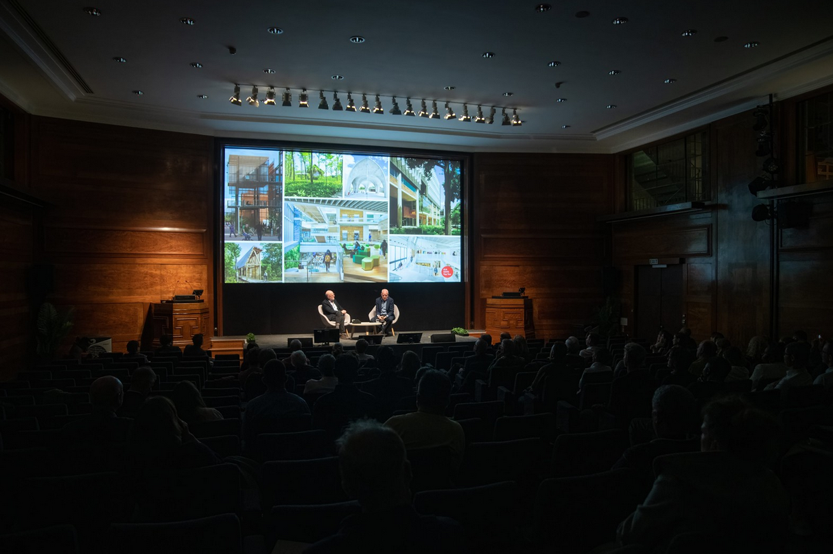 Two men are seated on stage on chairs, engaging in a discussion during a presentation or conference. Behind them, a large screen displays images of architectural designs and buildings. The room is dimly lit with an audience watching the event.