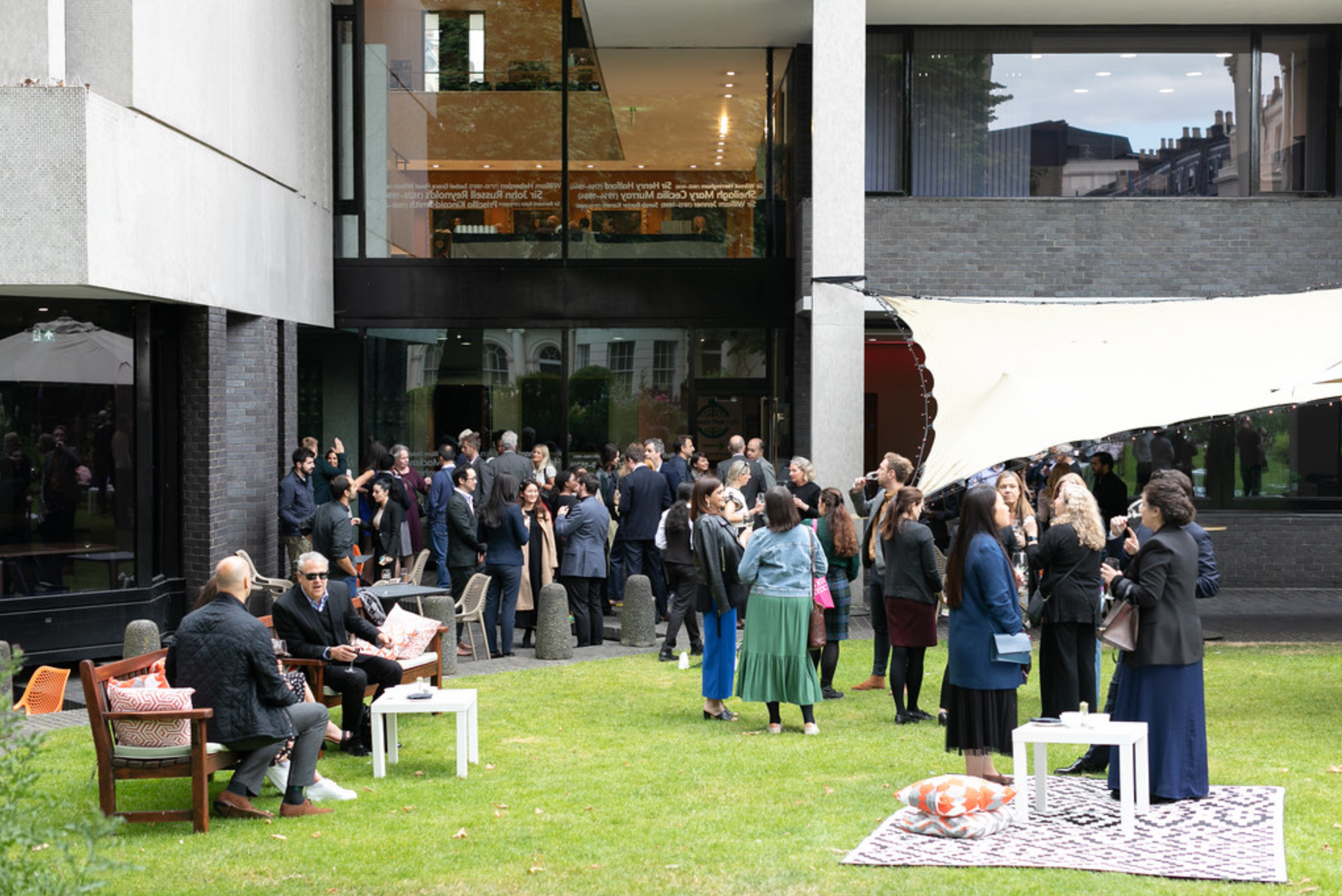 People gather outdoors at a social event on a grassy lawn in front of a modern building with large glass windows. Some are talking, while others are sitting on benches and chairs, and a few are standing near a white rug with pillows. The scene appears festive with groups chatting and enjoying drinks.