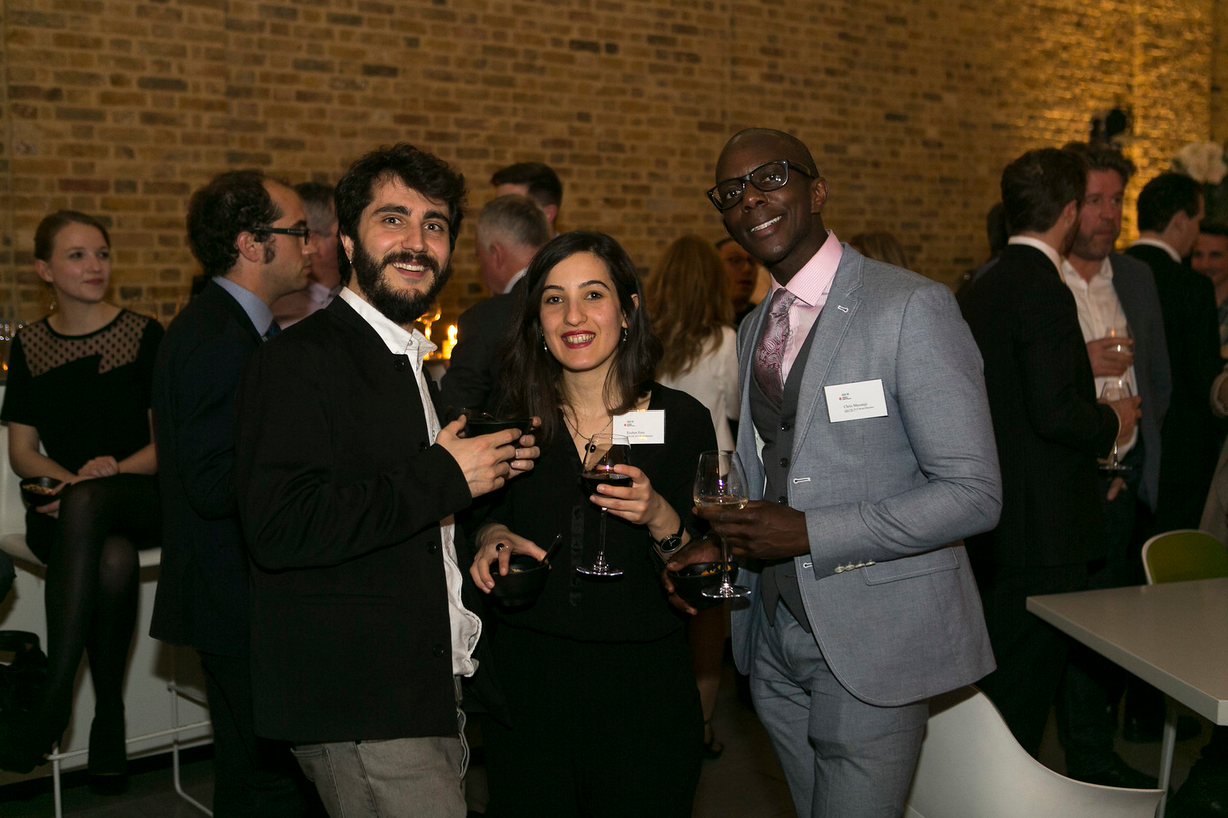 Three people smiling and holding glasses of red wine at a social event in a room with a brick wall, alongside other guests in the background.