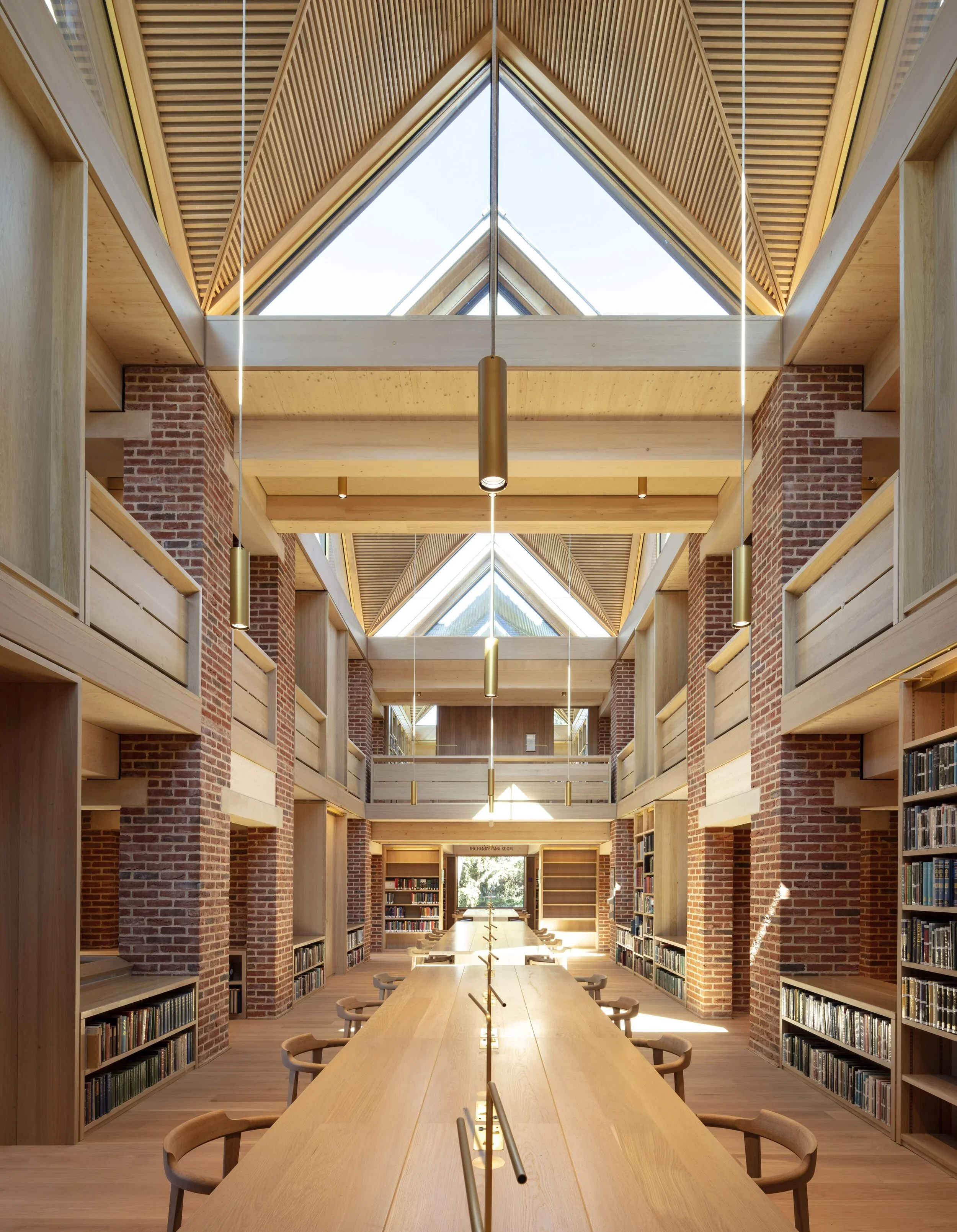 Interior of a modern library with a high vaulted ceiling, large triangular skylights, brick walls, and wooden shelving units. A long wooden table with chairs runs down the center, and bookshelves are filled with books along the sides.