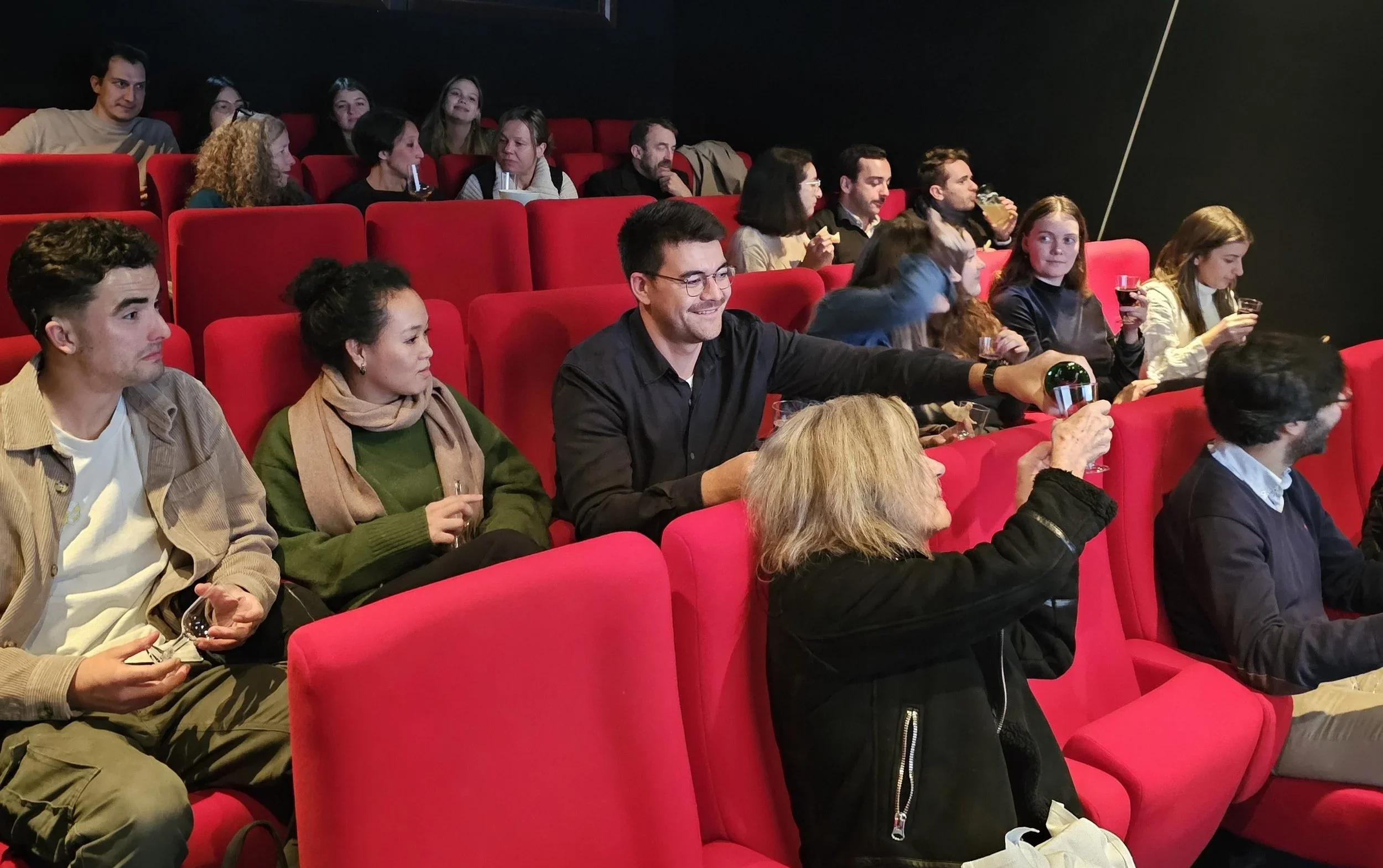 People sitting in a dark movie theater with red seats, some holding glasses of wine or drinks, engaging in conversation and watching a movie.