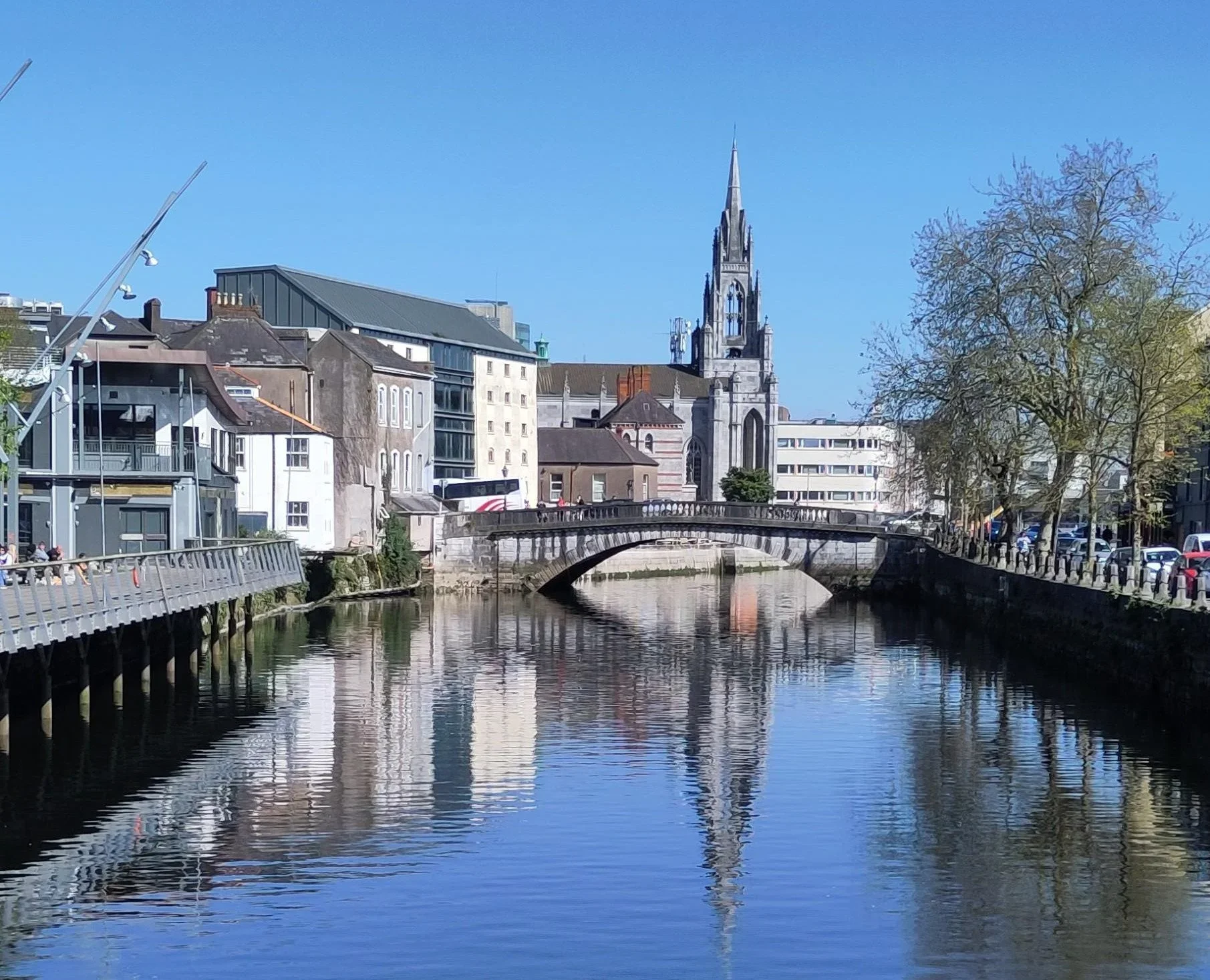 A cityscape with a river running through it, a stone bridge over the river, a church with a tall spire, trees on the right side, and modern buildings on the left, under a clear blue sky.