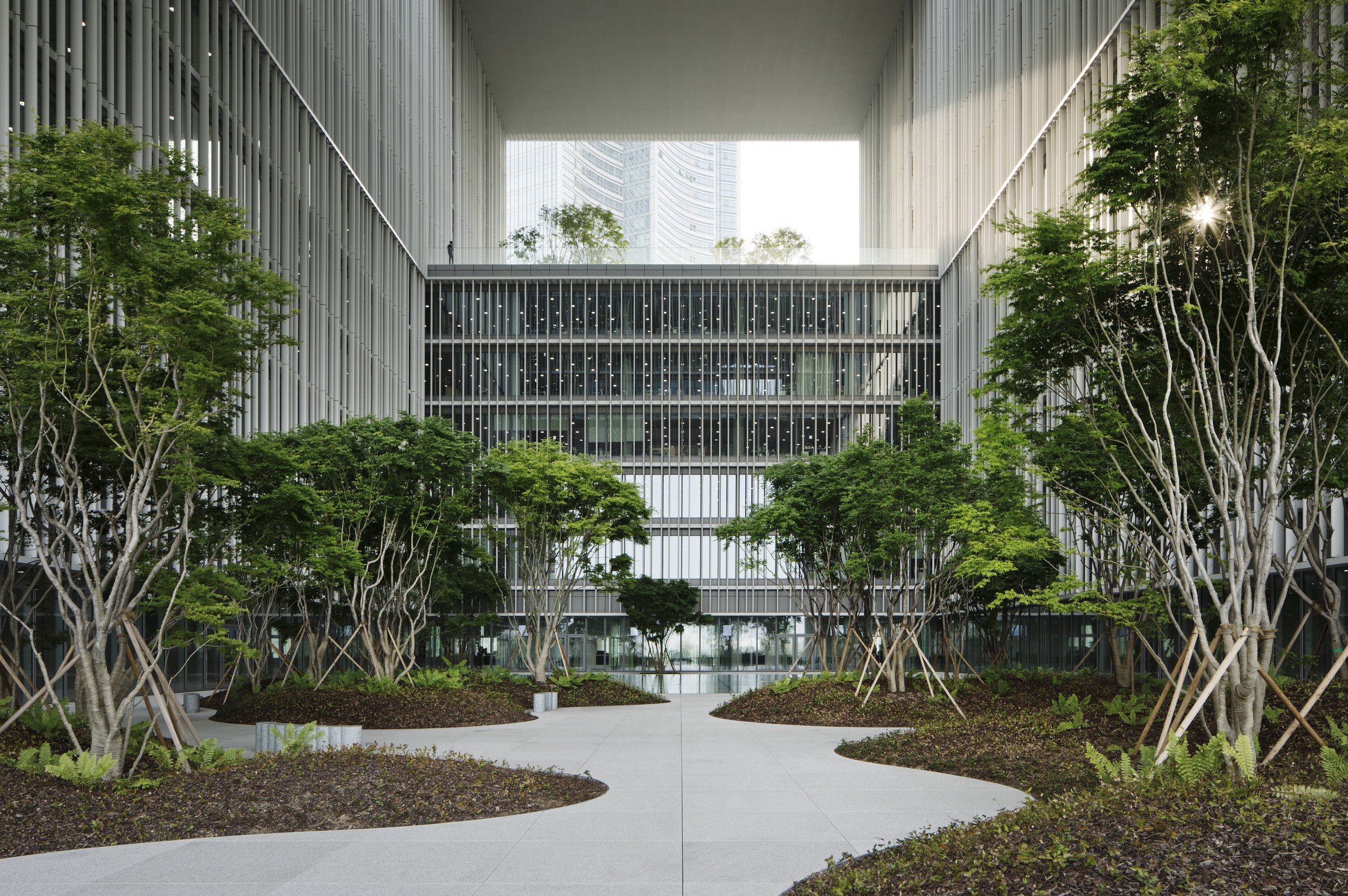 Modern urban courtyard with greenery, trees, and pathways surrounded by tall glass buildings with vertical metal slats.