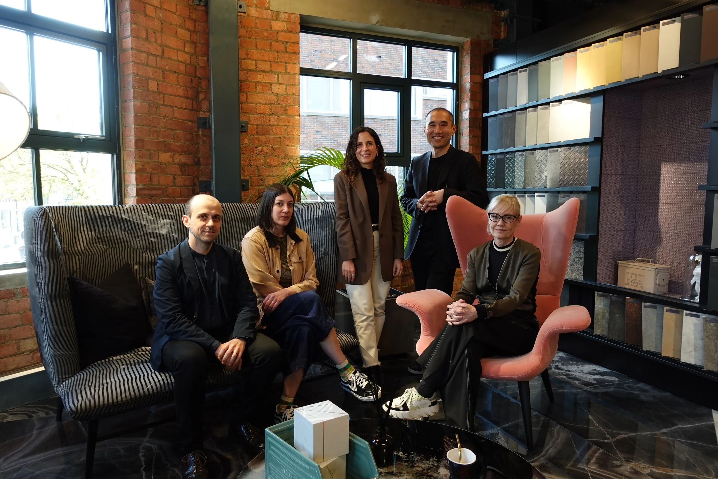Group of five diverse professionals in a modern office lounge, with brick walls, large windows, and stylish furniture, smiling for the photo.