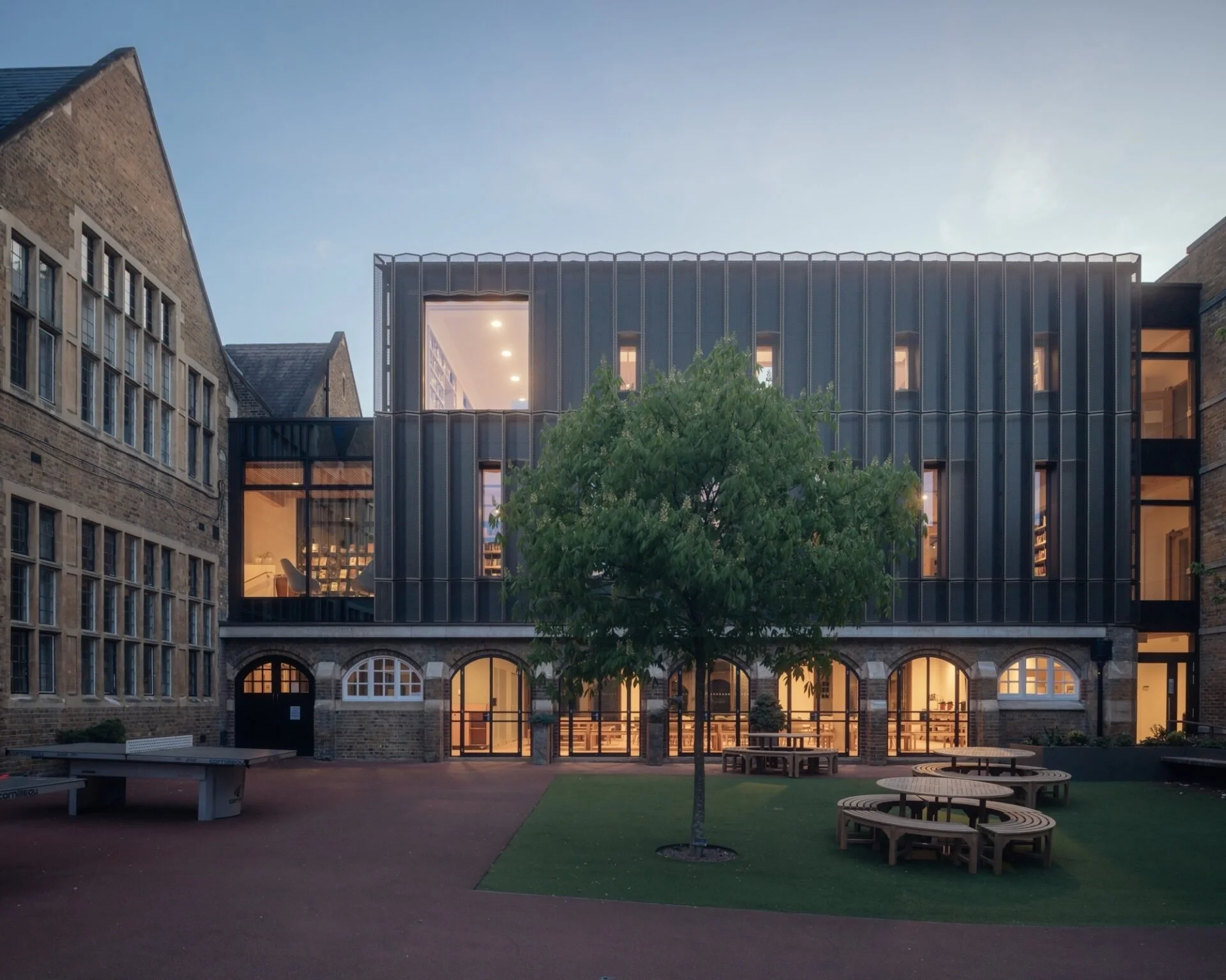 Modern building with glass windows and brick facade, surrounded by a small courtyard with benches and a tree, during evening with interior lights on.