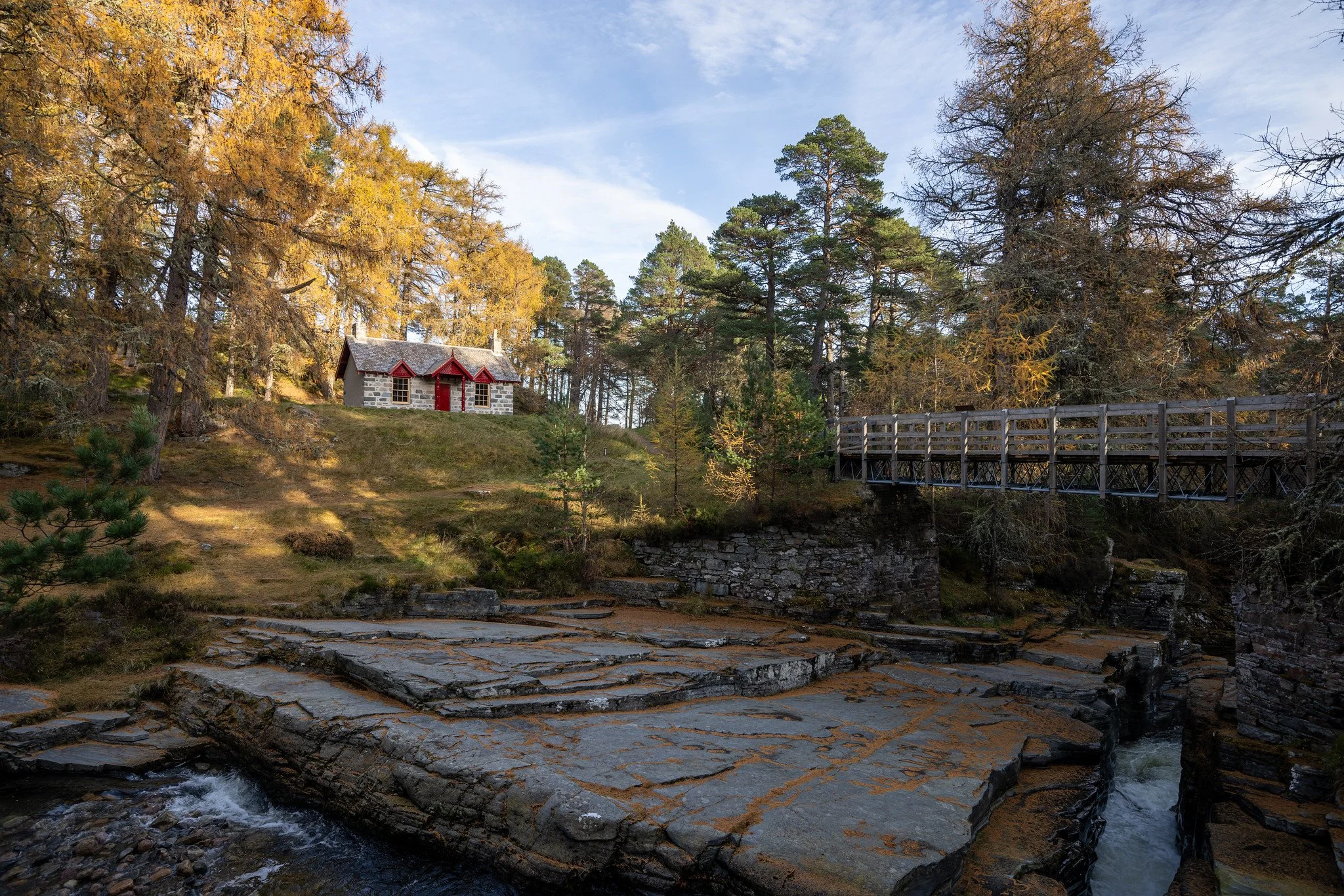 A small stone house with red trim on a grassy hill surrounded by trees with changing leaves, a wooden bridge crossing a rocky stream, and a clear blue sky overhead.