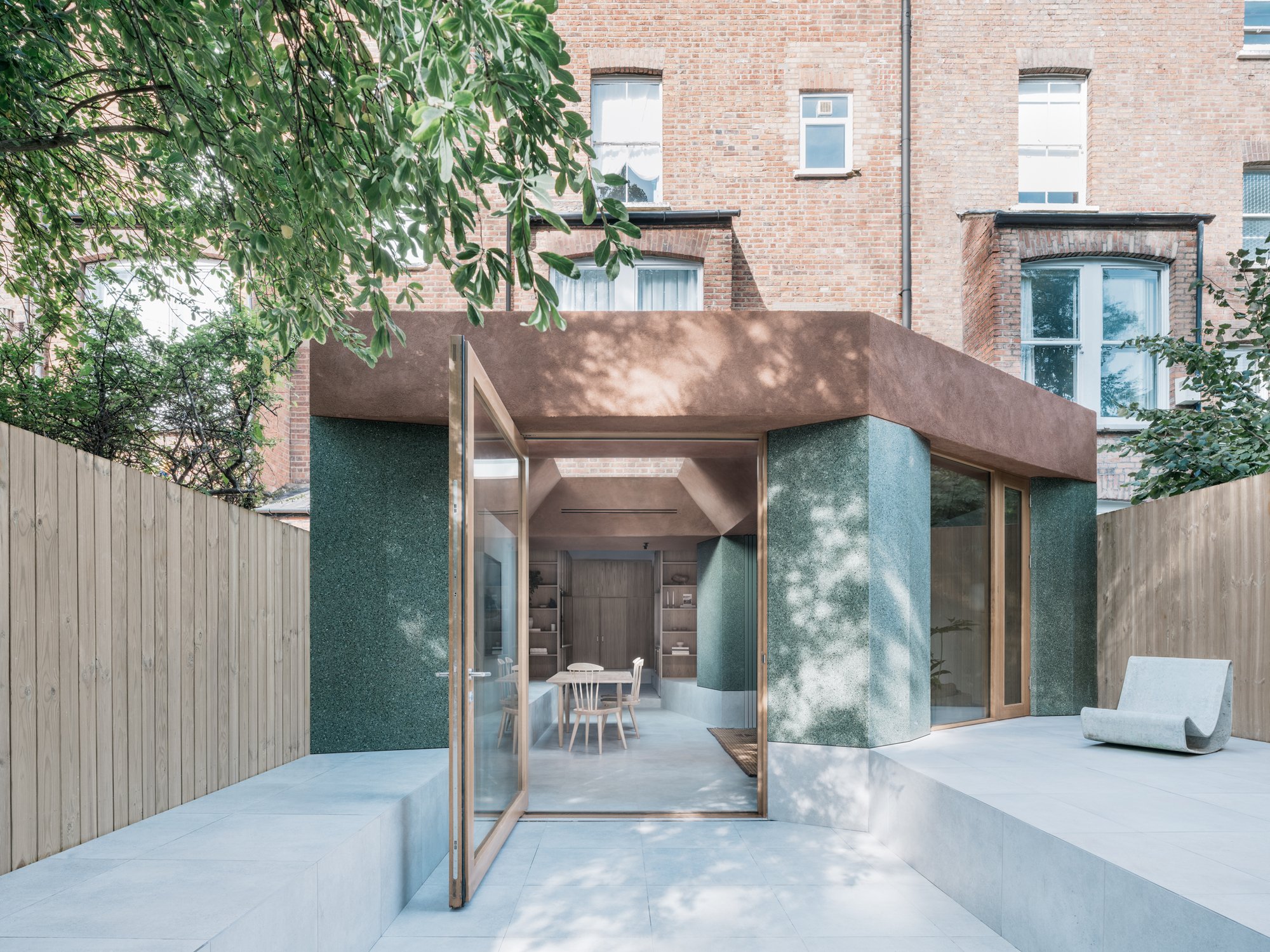 Modern backyard with a patio, wooden fence, and a small, enclosed garden room with large glass doors, beige walls, and an integrated interior dining area visible inside. A curved concrete bench is on the patio, and trees provide shade.