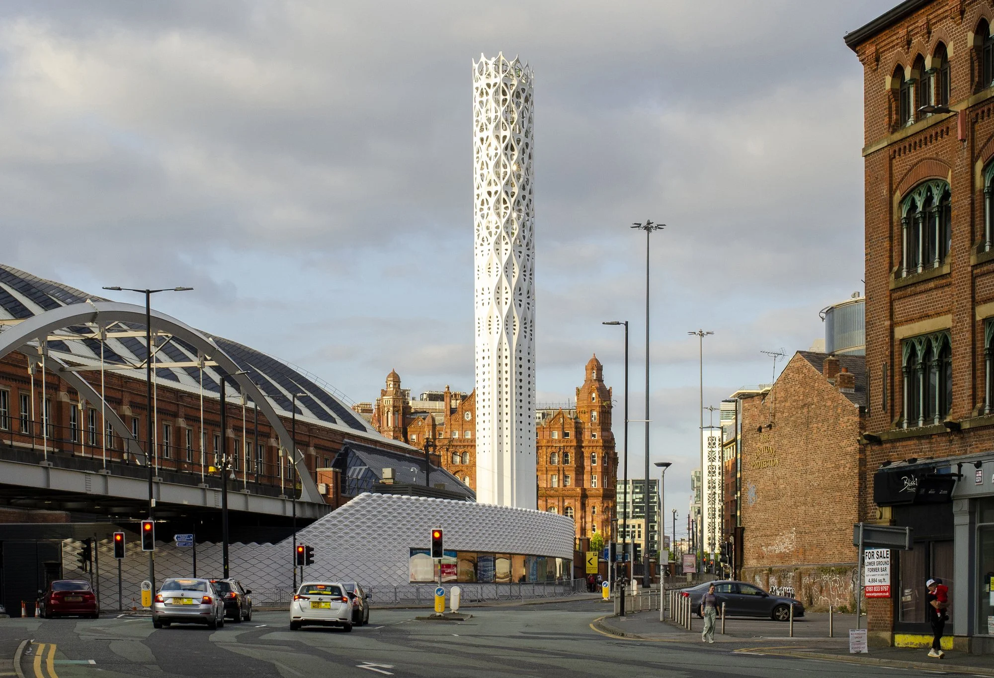 A city street scene with a modern white tower structure, historic brick buildings, cars on the road, and pedestrians, under a partly cloudy sky.