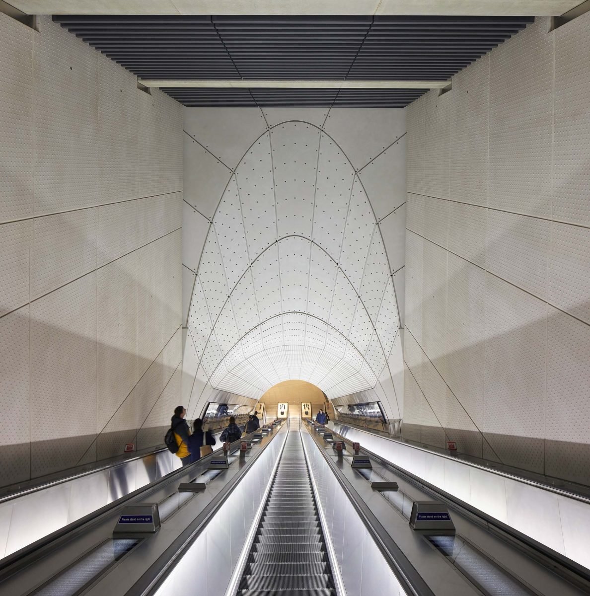 An underground subway station with escalators, people riding, and a modern, geometric ceiling design.