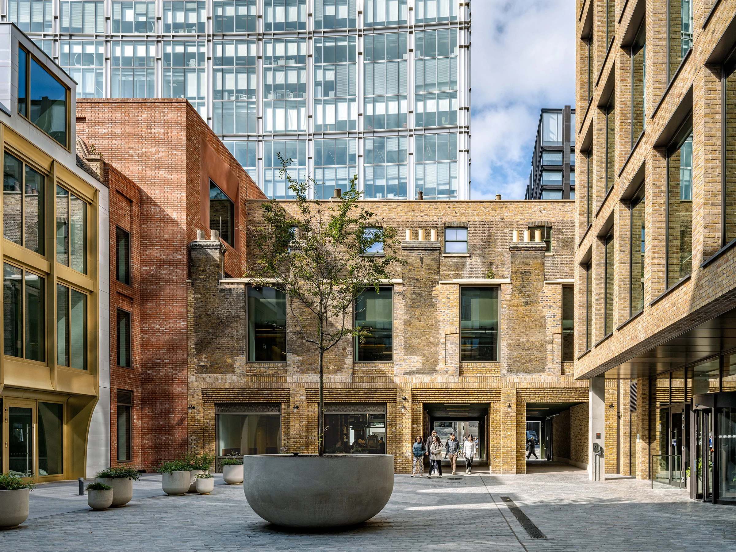 Urban courtyard surrounded by modern and historic brick and glass buildings with pedestrians walking through.