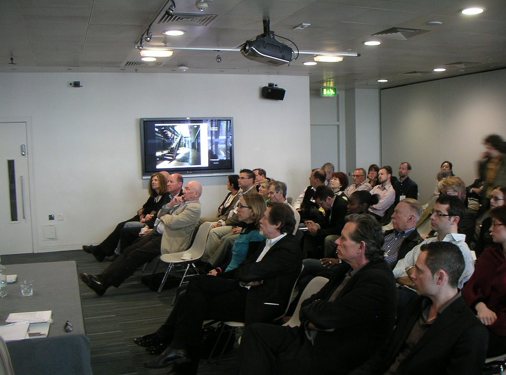 A group of people seated and watching a presentation in a conference room with a television screen on the wall.
