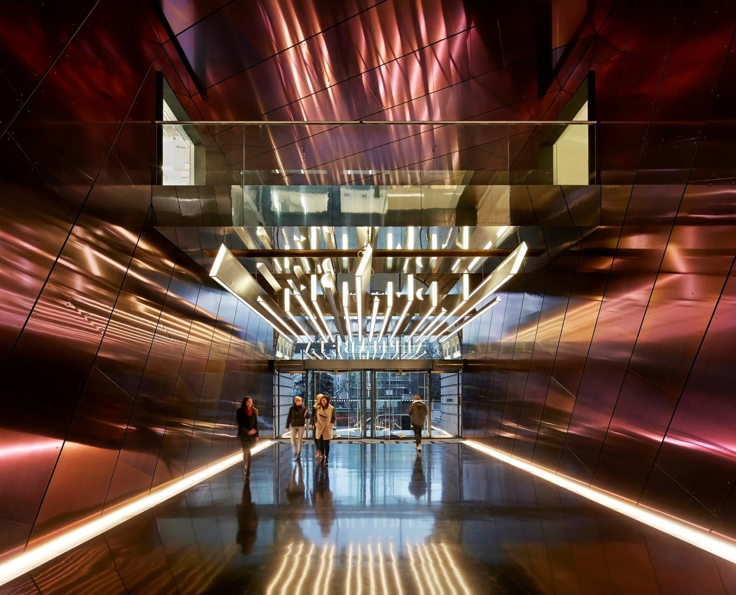 Interior of a modern building with metallic walls, illuminated geometric hanging light fixtures, and a glass entrance area with people walking inside.
