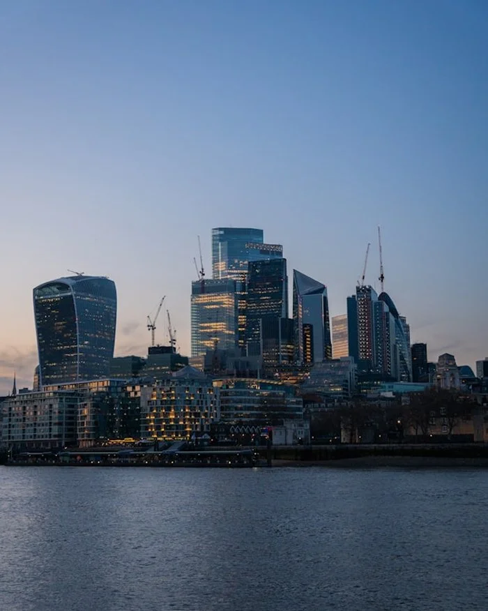 Skyline of modern skyscrapers near a river, with visible construction cranes and a partly cloudy sky.