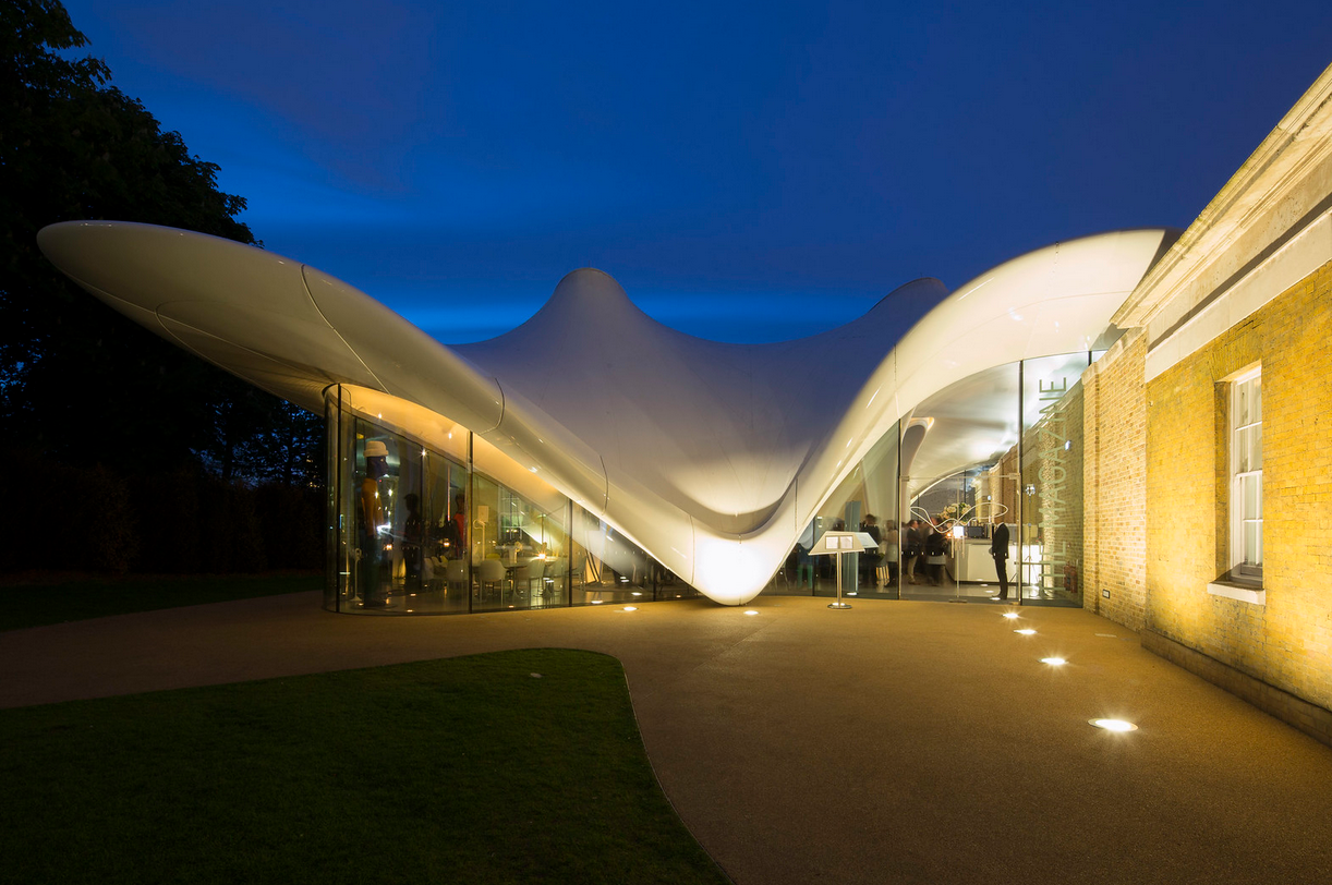 Night view of the innovative design of the LVMH Tower, featuring a curving white structure with glass walls, illuminated from within, adjacent to an older brick building.