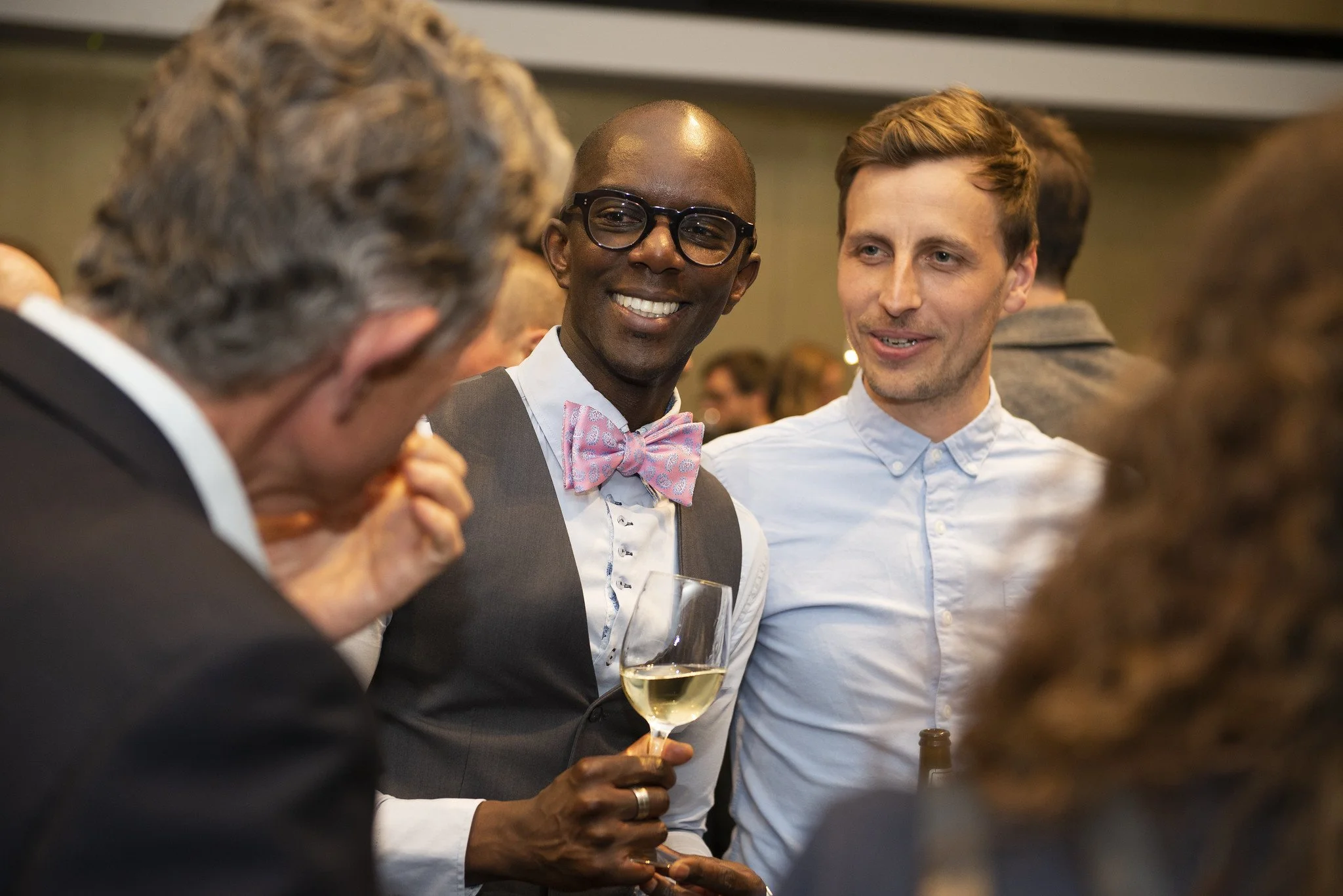 A group of people at a social event, including a man in a suit with glasses and a pink bow tie holding a glass of white wine, engaging in conversation with others in a crowded room.