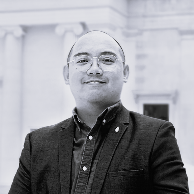 A man with glasses wearing a dark blazer and collared shirt, smiling indoors with classical architecture in the background.