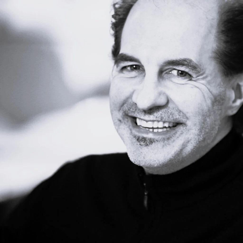 Black-and-white close-up of a smiling man with dark hair and a light beard, wearing a black top.