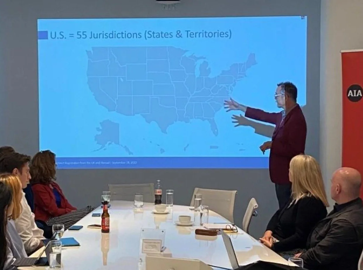 A man is giving a presentation about U.S. jurisdictions, with a map of the United States projected on a screen. Several people are seated at a table, watching and listening.