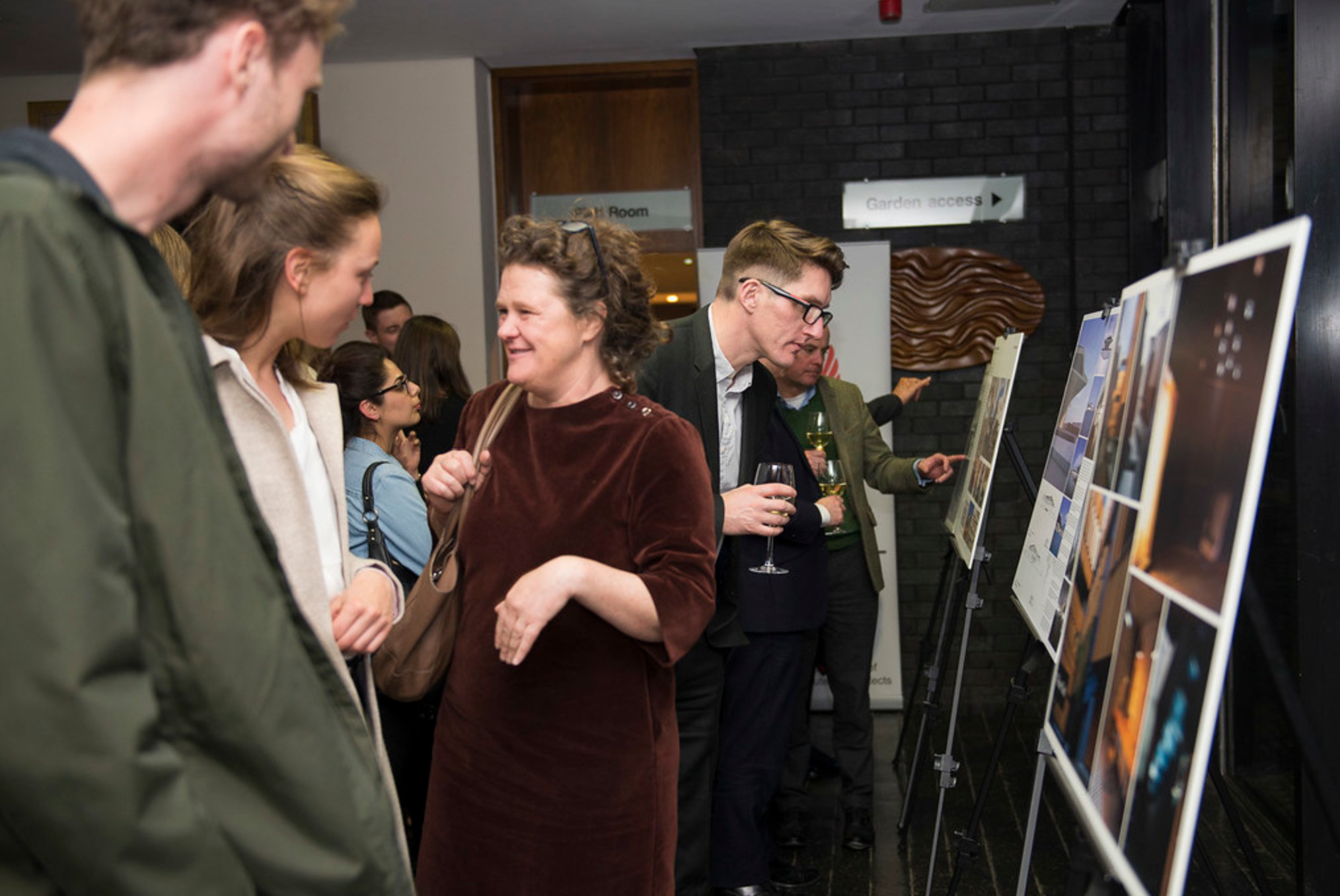 People viewing art photos on display boards at an indoor gallery event, some holding glasses of wine.
