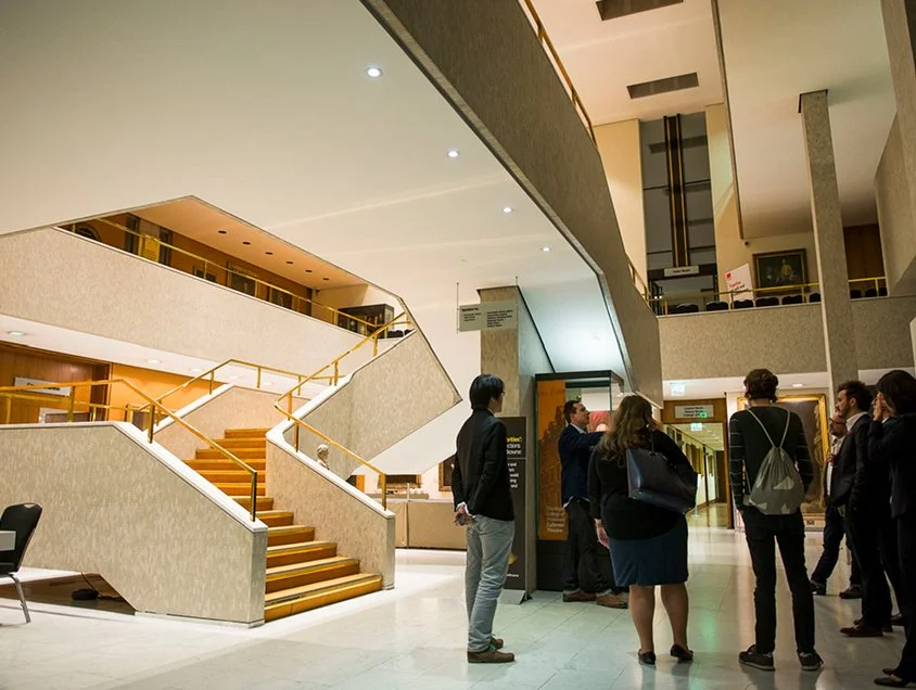 People standing and talking in the lobby of a modern building with staircase and upper levels visible.