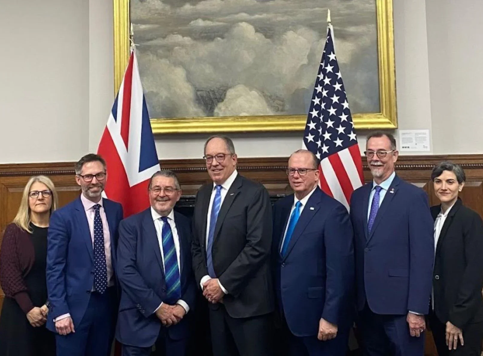 Group of seven people, five men and two women, standing in front of British and American flags, with a framed painting behind them.