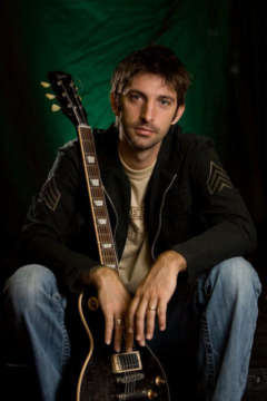 A man with short brown hair and a beard, sitting and holding a black electric guitar in front of a green and black background.