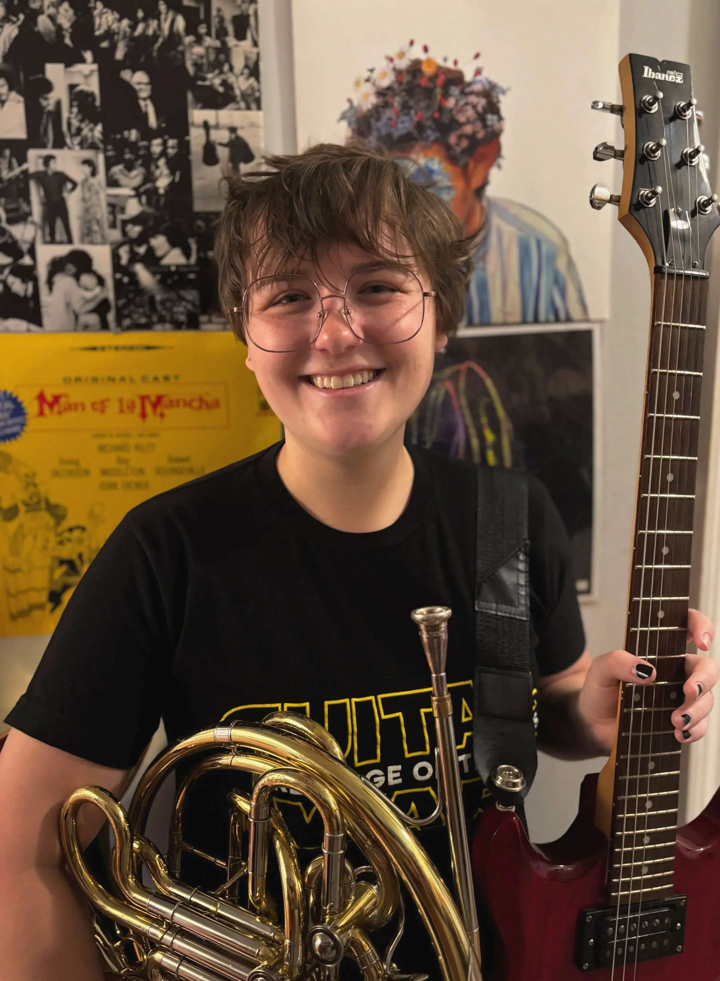 A person with short, curly brown hair and glasses smiling while holding a French horn in one hand and a red electric guitar in the other. They are wearing a black t-shirt with yellow and white designs. Behind them are various posters and artwork on the wall, including a black and white collage and a colorful portrait.