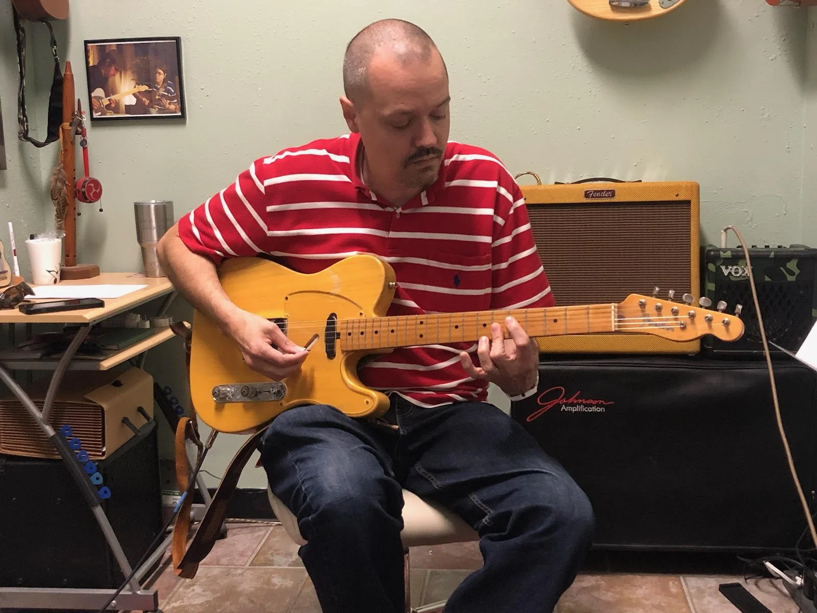 A man with a shaved head wearing a red and white striped shirt playing an electric guitar in a room with musical equipment and a wall decorated with a framed picture.