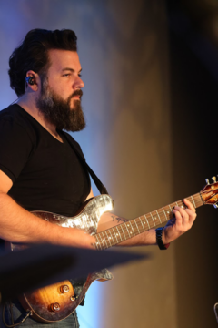 A man with dark hair and a beard playing an electric guitar on stage.