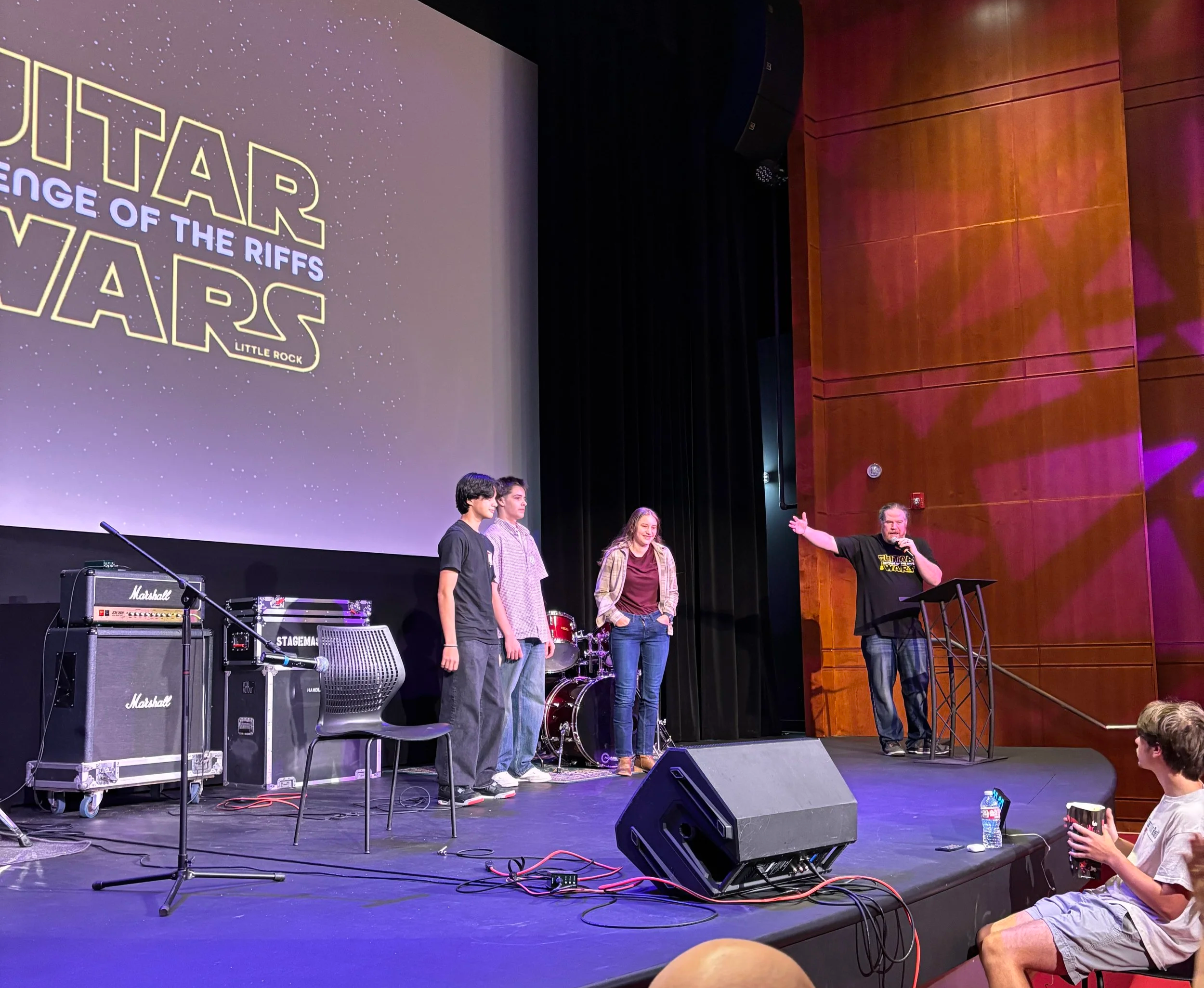 People on stage during an event with a large screen in the background displaying the Star Wars logo and the title 'Rise of the Riffs' Little Rock. There are musical instruments such as amplifiers, drums, and microphones set up on stage. A man is speaking at a podium with his arm extended, and three others are standing nearby. Audience members are seated, including a person in the front holding a cup and a water bottle.