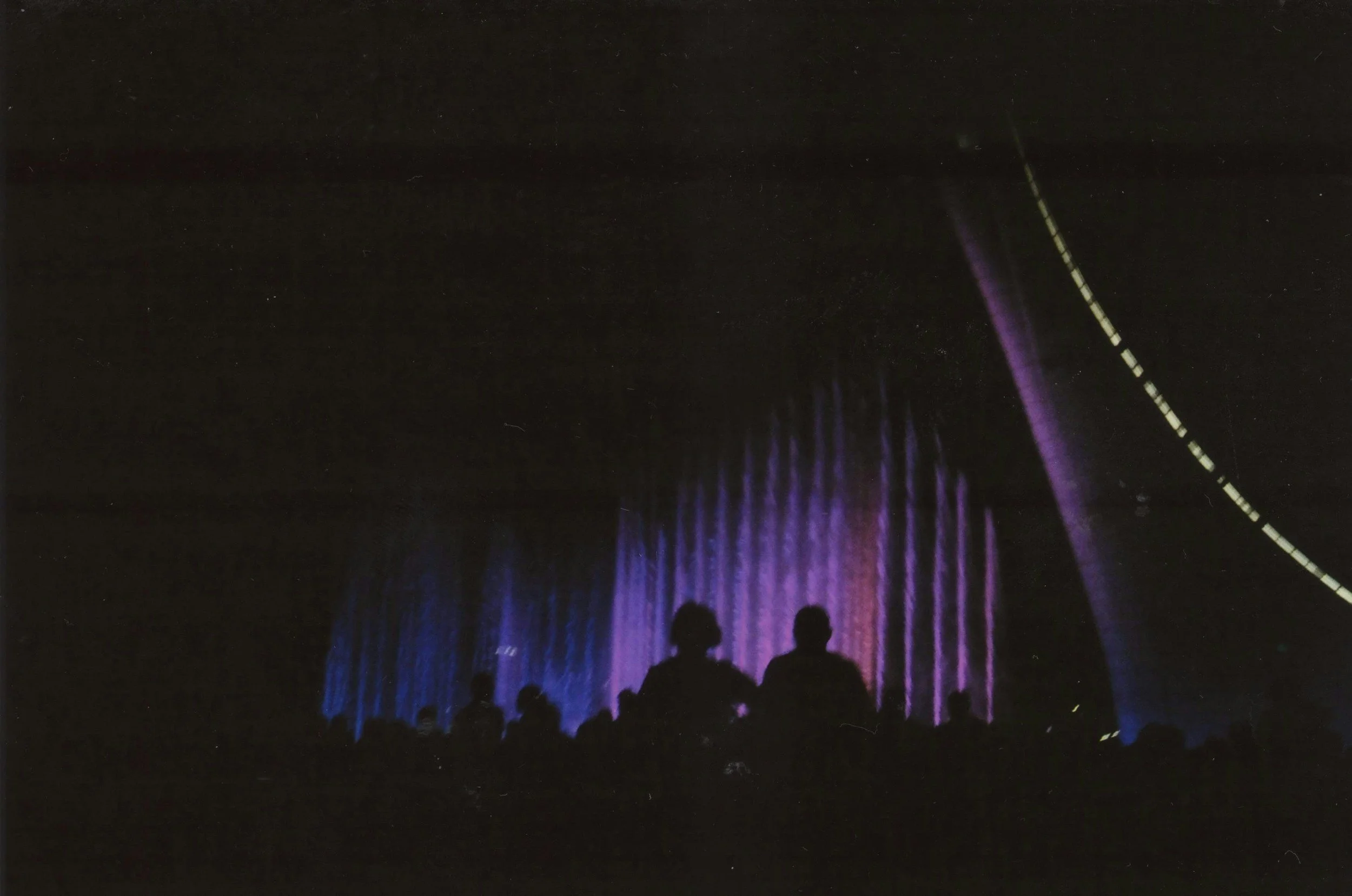 Silhouettes of people watching a colorful, illuminated fountain display at night.