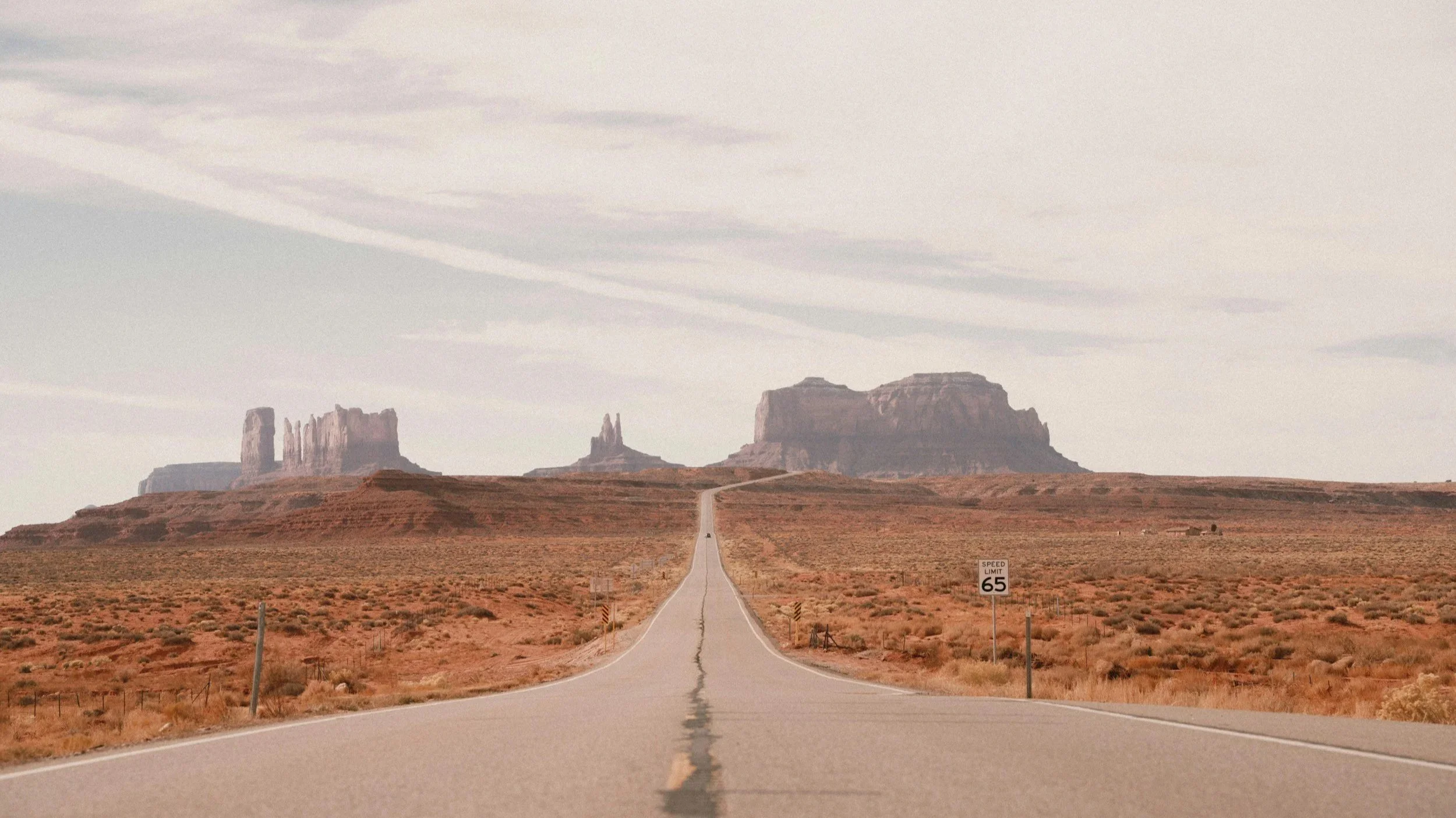 A straight road in a desert landscape with rock formations in the distance and a cloudy sky.
