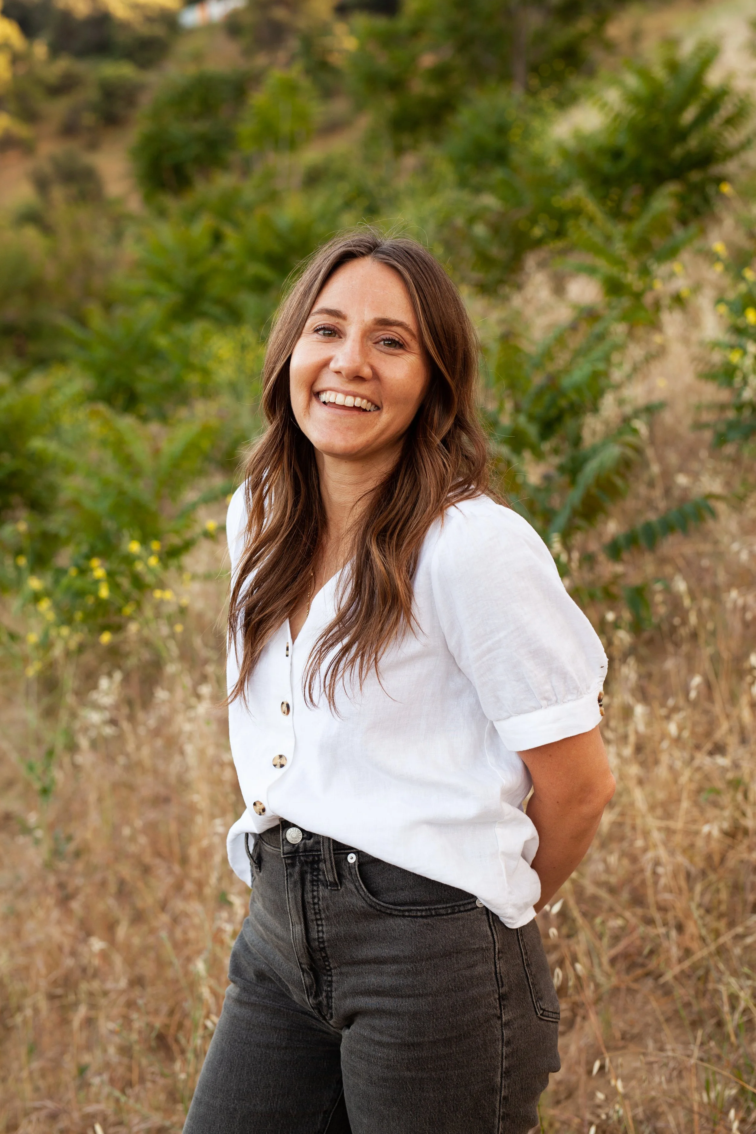 A woman with long brown hair smiling outdoors among green plants and dry grass.