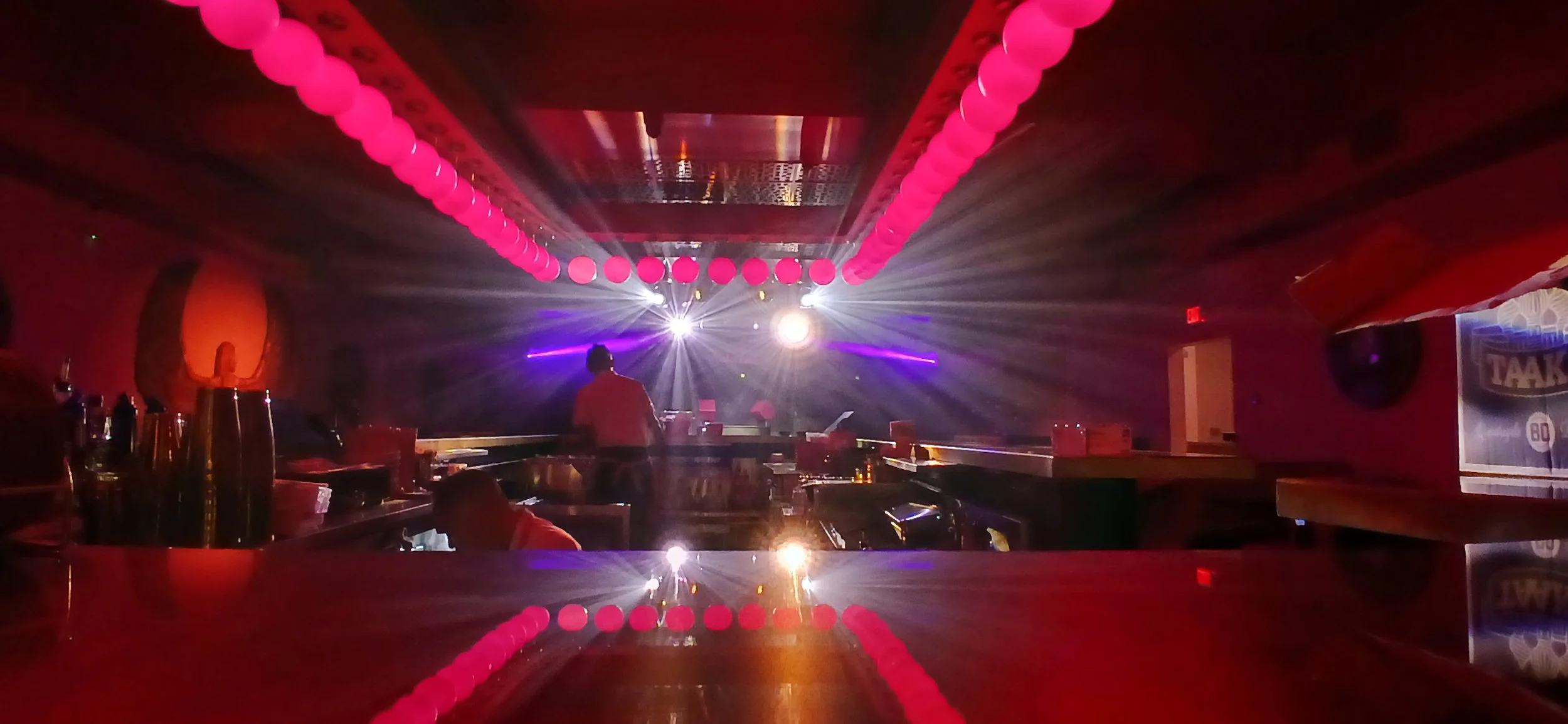 Club or concert venue with pink hanging lanterns, dance floor, DJ booth, and light beams from the DJ or stage area.