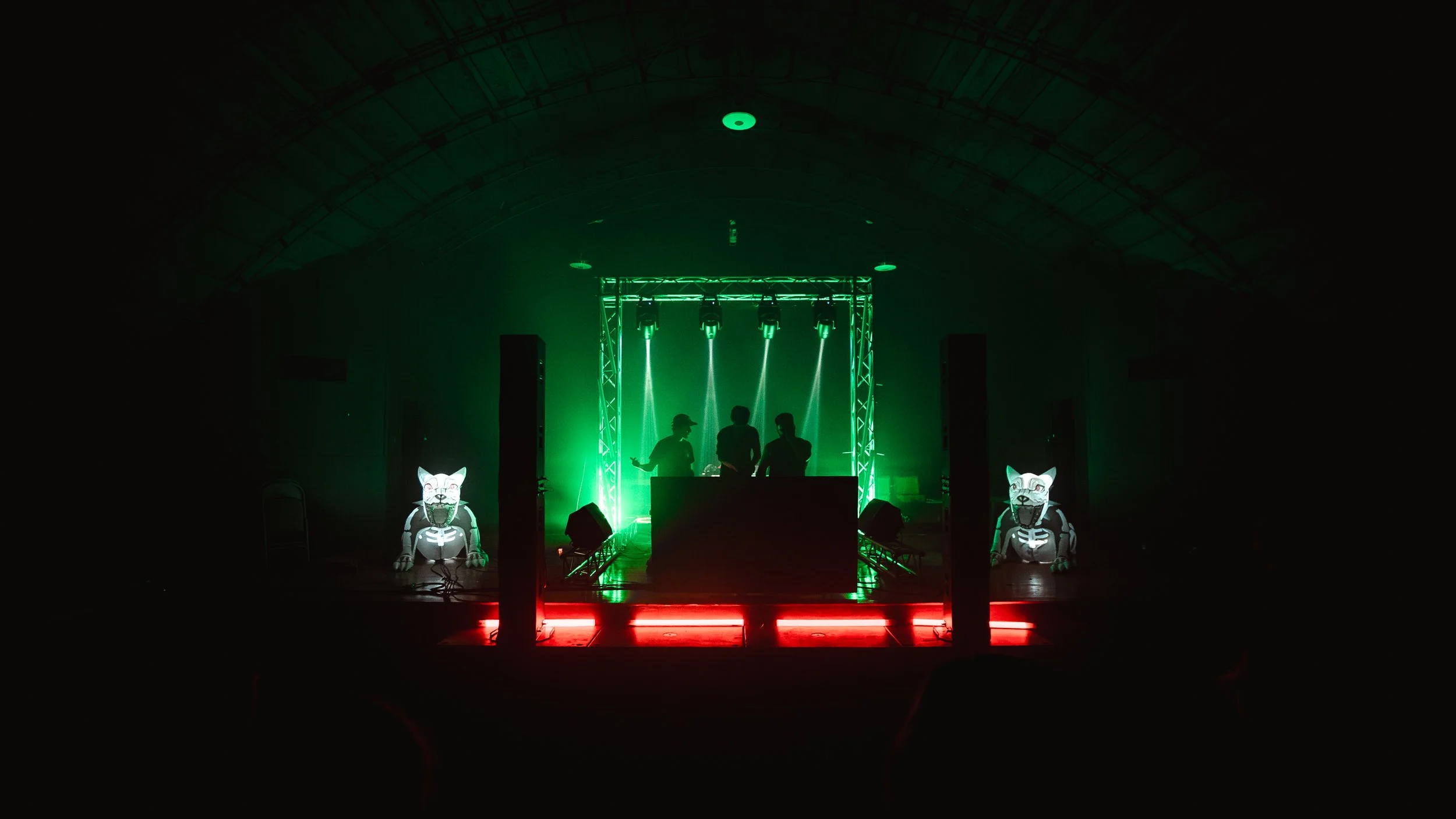 Dark indoor nightclub scene with green and red lighting, three silhouetted DJs or performers on stage behind a DJ booth, two large illuminated wolf or dog figures on the floor on either side of the stage.