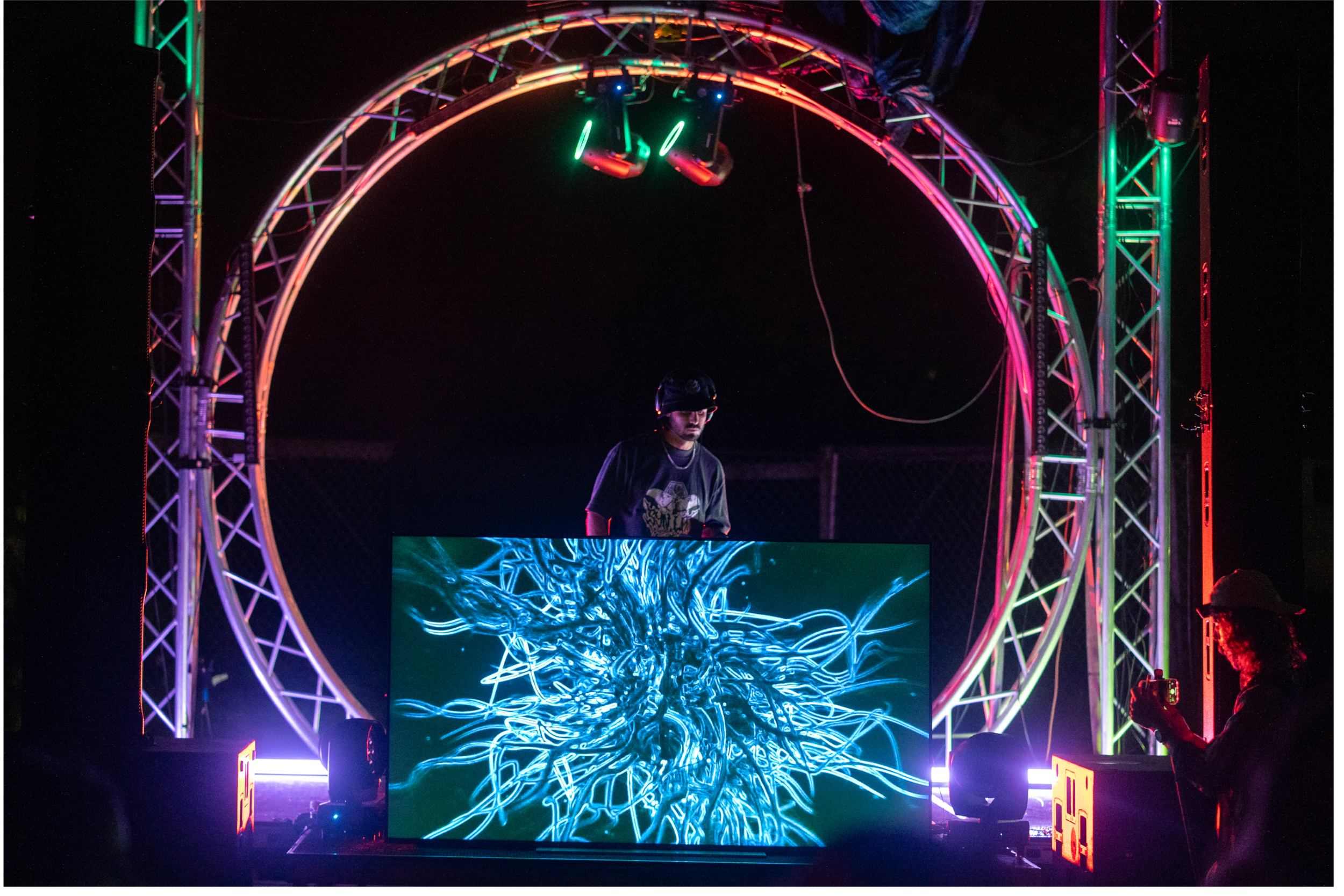 A DJ performs at an electronic music event, standing behind a large digital screen displaying a blue, abstract, tree-like digital art. The DJ is under a circular metal frame with colorful neon lights, with two spotlights or moving head lights mounted above. A person in the foreground on the right side is taking a photo or video with a smartphone.