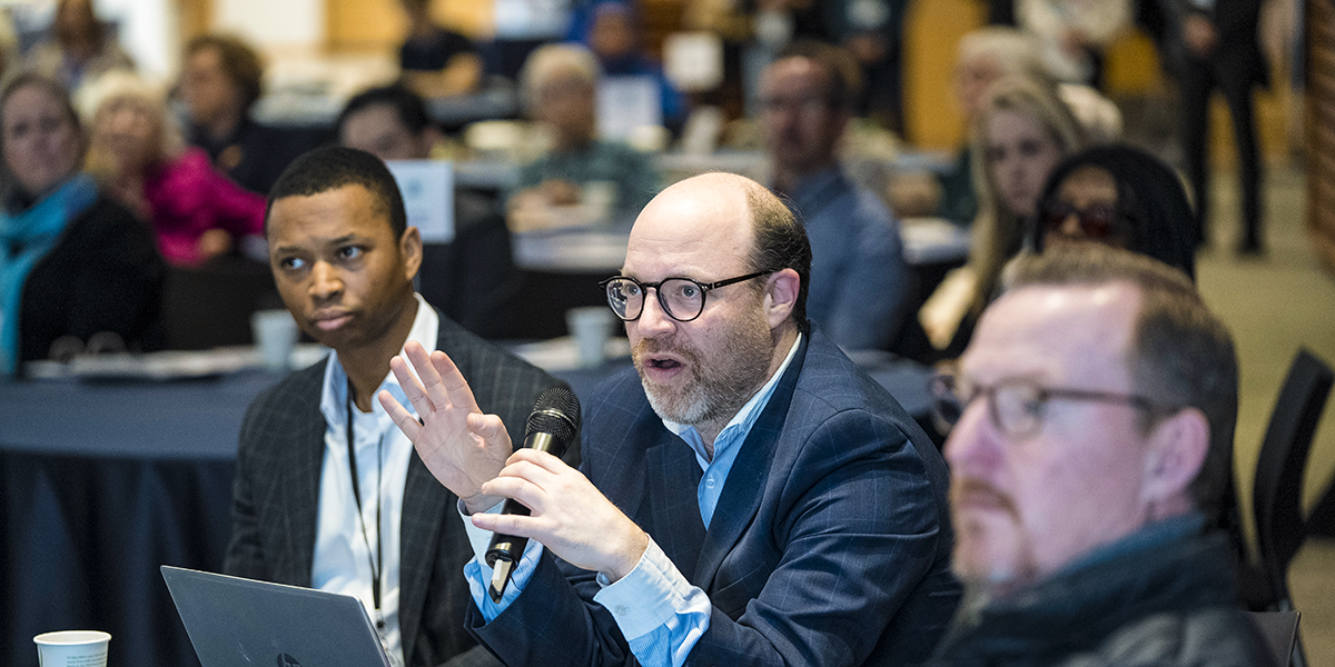 A conference attendee speaks into a handheld microphone during a conference Q&A session. He is surrounded by fellow attendees seen in the foreground and background.