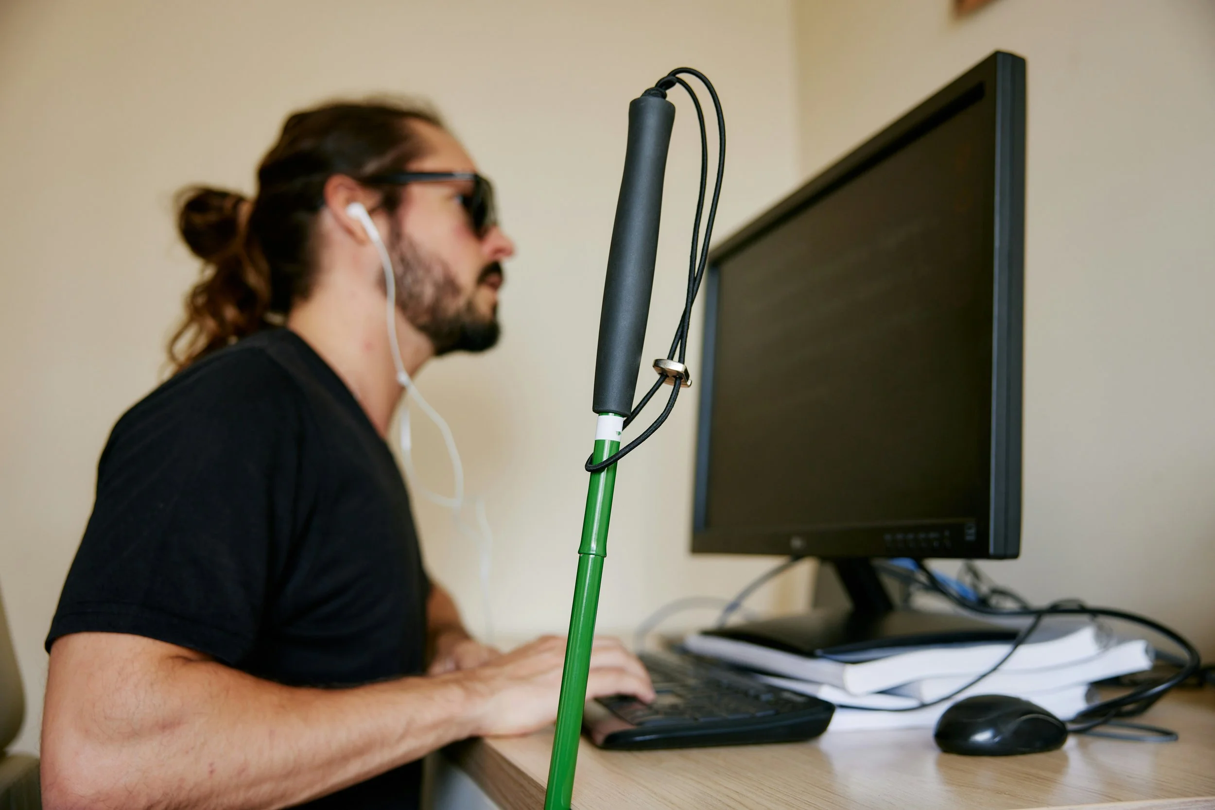 A man living with blindness sitting in front of a computer with his hands on a keyboard and wearing headphones. His cane is shown in focus in the foreground.