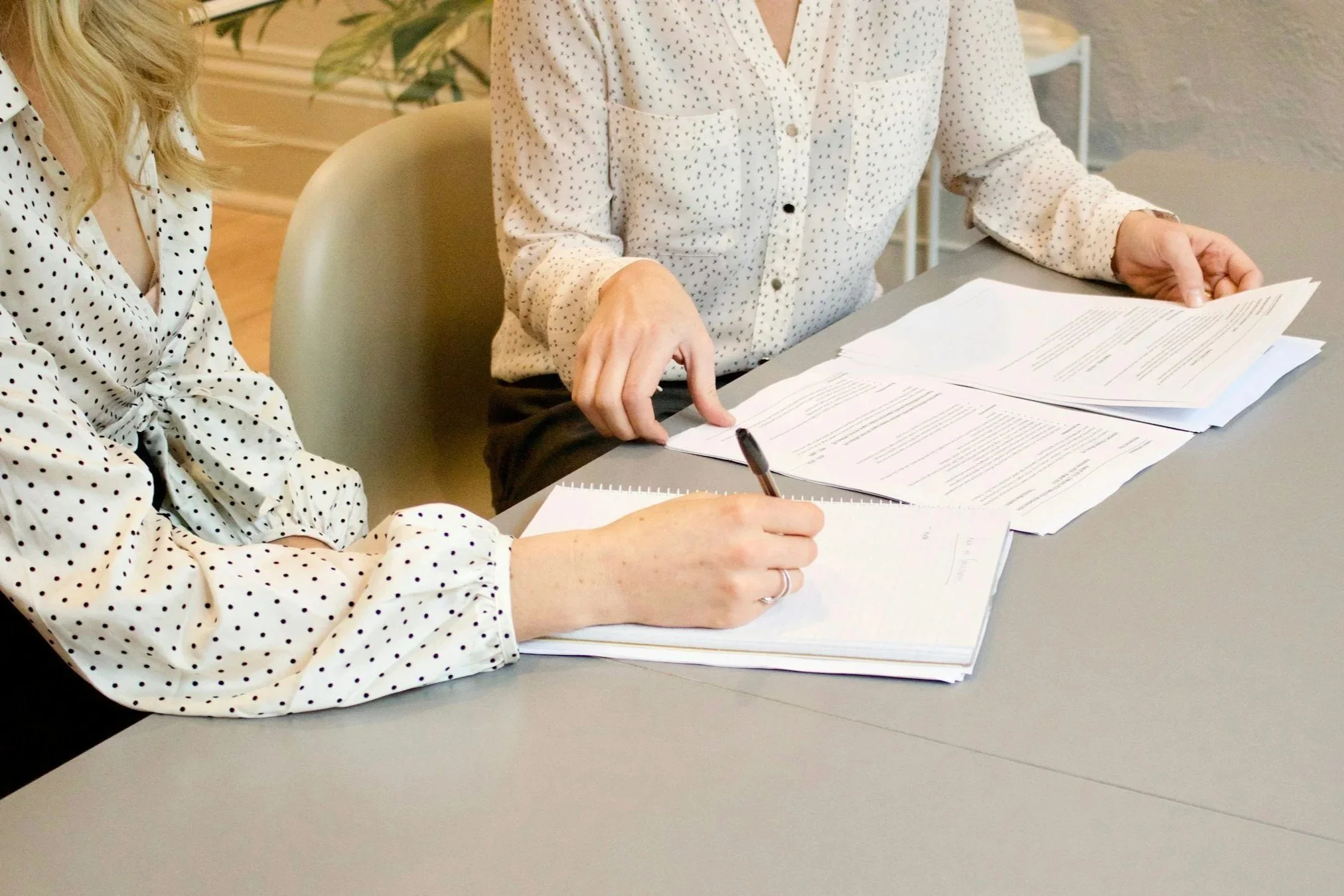 Two women in polka dot shirts sitting at a table, reviewing and taking notes on printed documents.