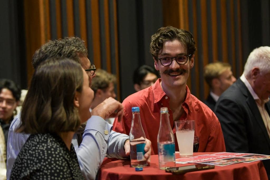 A group of people sitting and chatting at a round table during a social event with wood-paneled walls in the background.