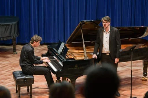A pianist playing a grand piano and a singer performing on stage in front of an audience.