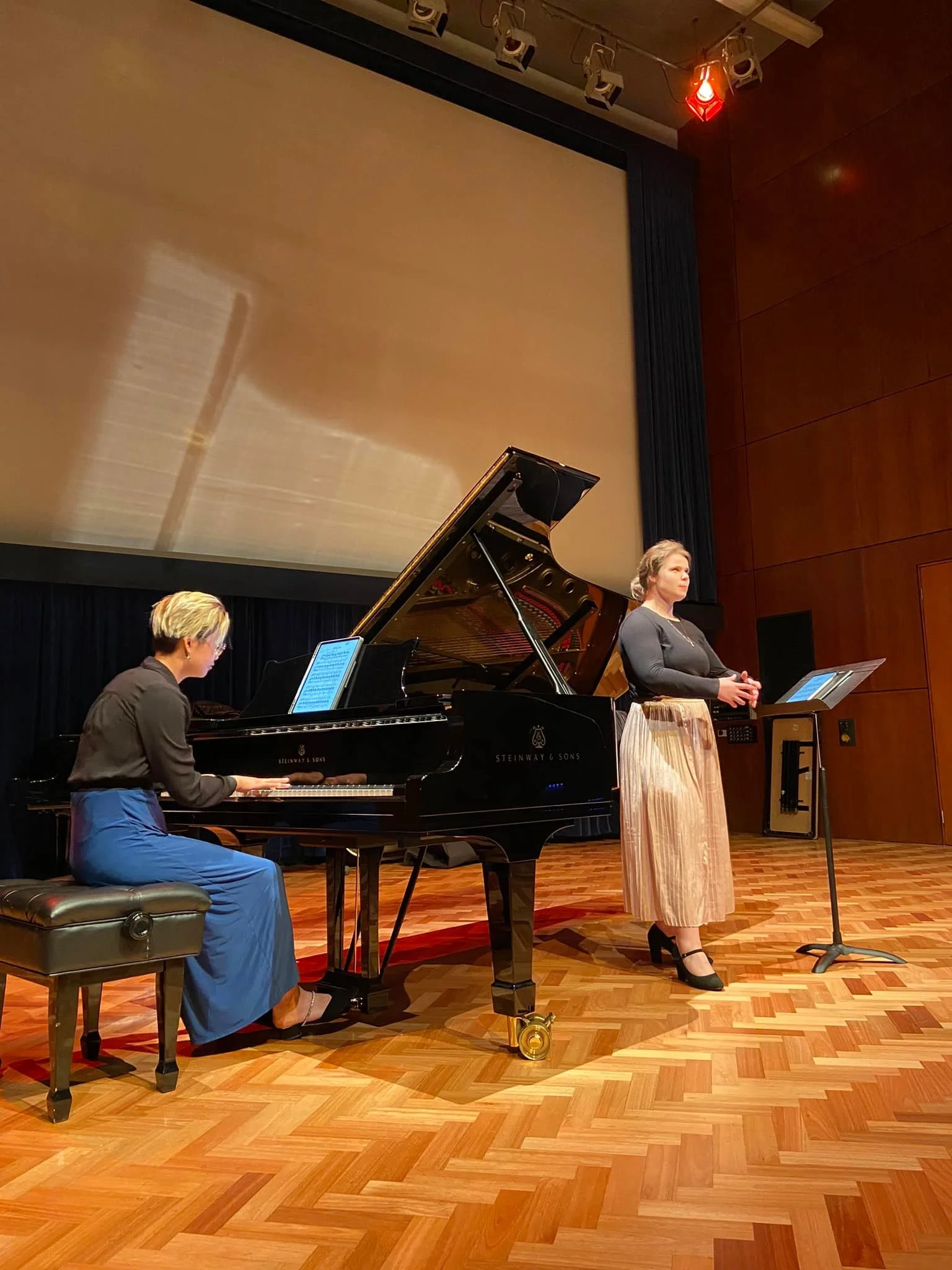 A woman stands on stage next to a music stand, singing or speaking, while another woman plays a grand piano on her left.