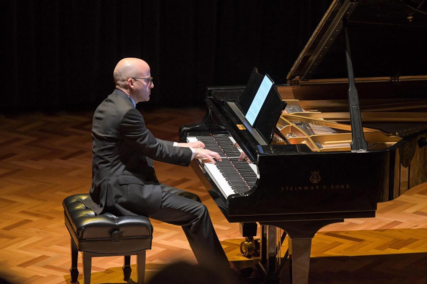 A man in a formal suit playing a grand piano on a wooden stage.