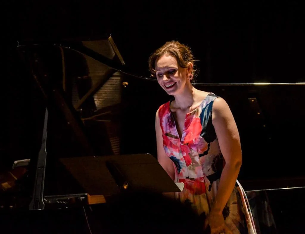 A woman playing the piano on stage, smiling, with a black grand piano in a dimly lit setting.