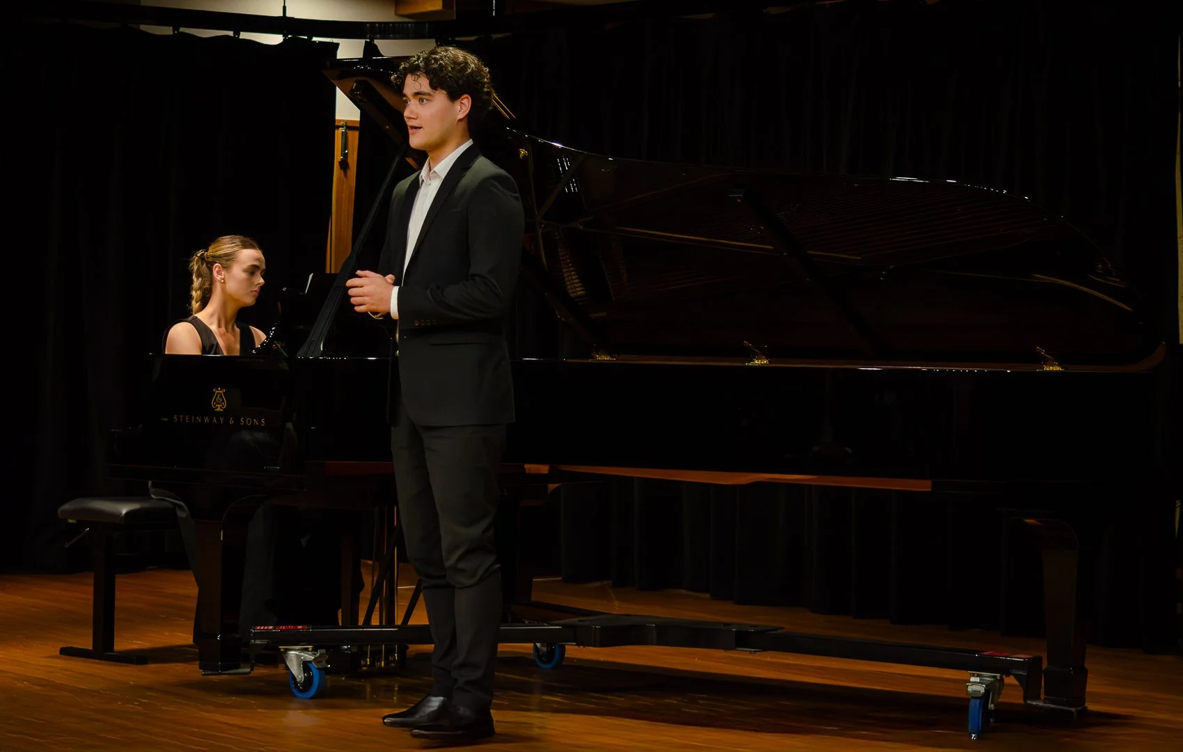 Young man in a tuxedo performing on stage with a grand piano in the background, with a woman playing the piano beside him.