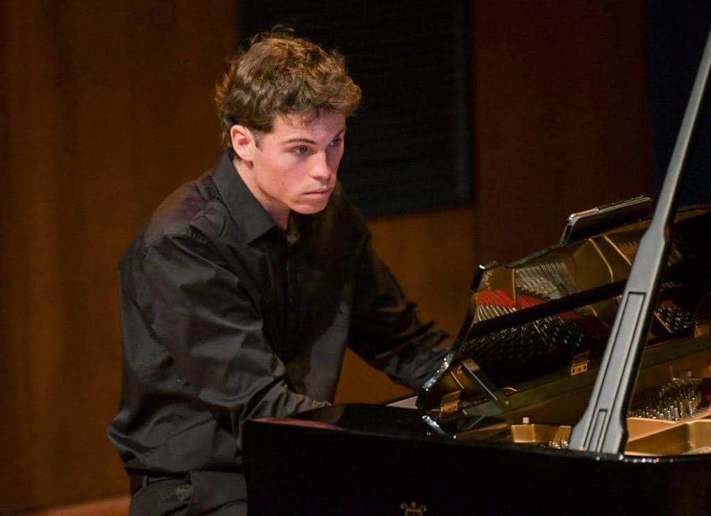 A young man with brown curly hair playing a black grand piano in a wooden-paneled room.