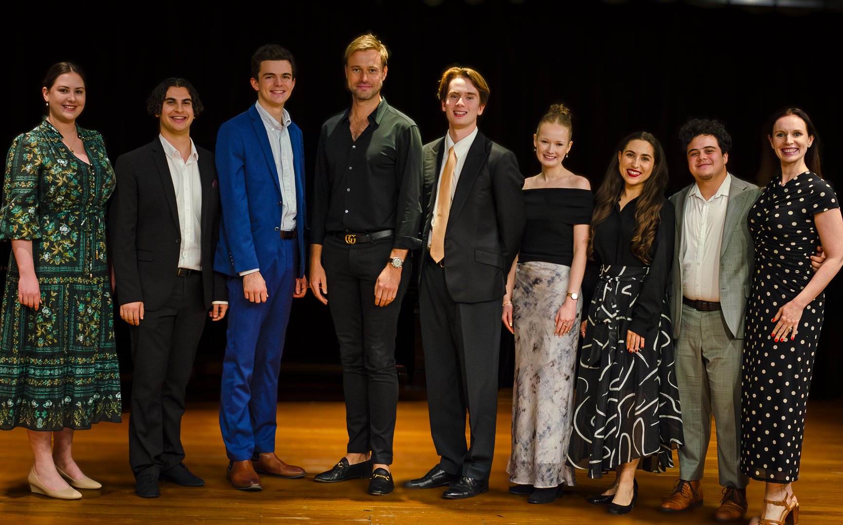 Group of nine people standing on stage with a dark background, dressed in formal and semi-formal attire, smiling at the camera.