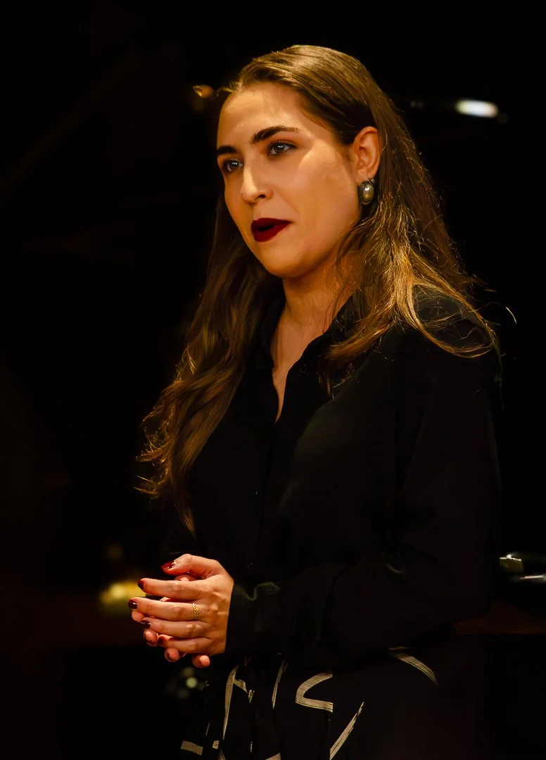 A woman with long, wavy brown hair, wearing a black outfit and pearl earrings, standing with her hands clasped, speaking or giving a speech in a dimly lit setting.
