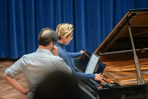 Two people at a piano, one is sitting and the other standing, looking at sheet music.